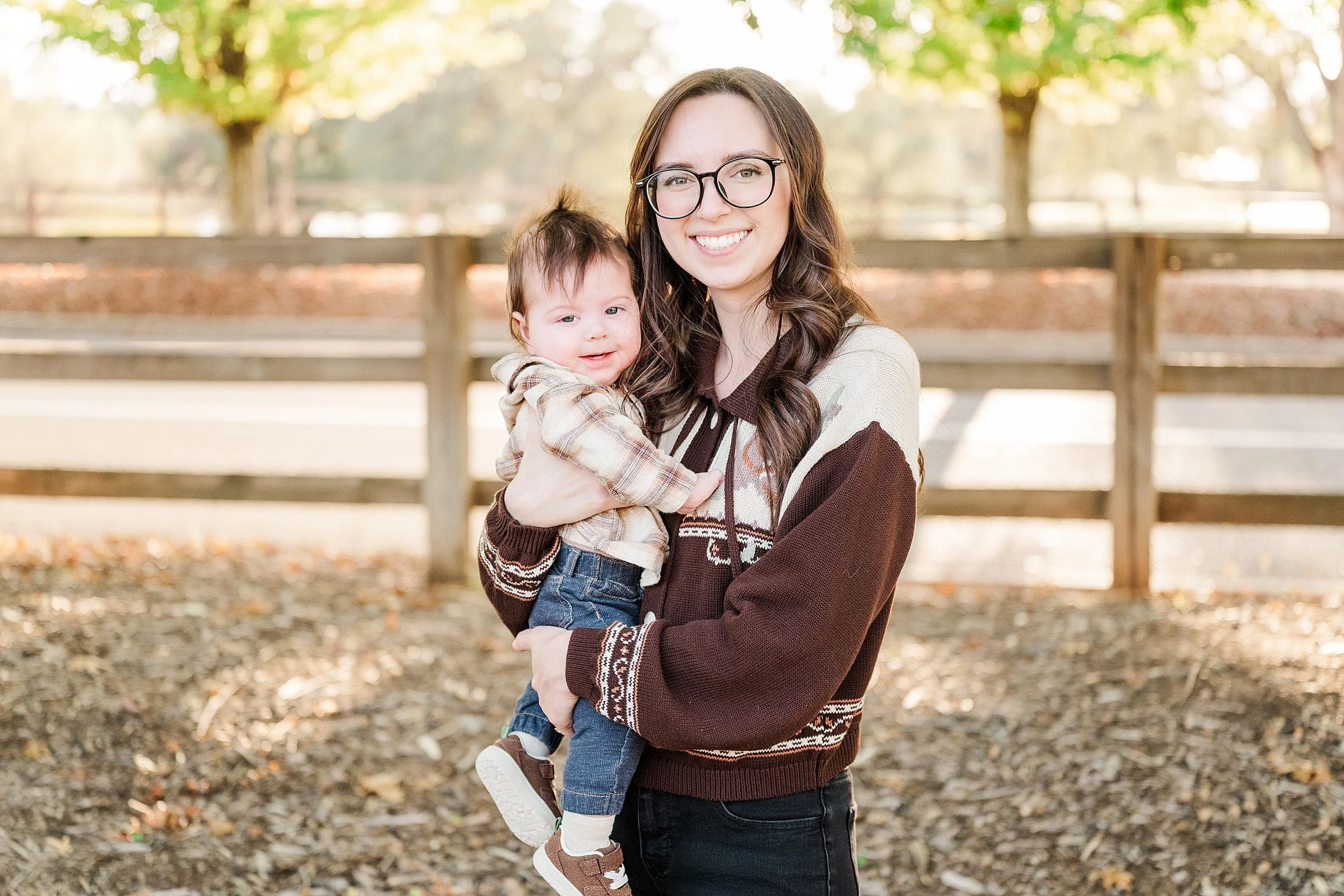 A smiling woman in glasses holds a toddler outdoors beside a wooden fence, with sunlit autumn trees behind them; the portrait feels warm and joyful.