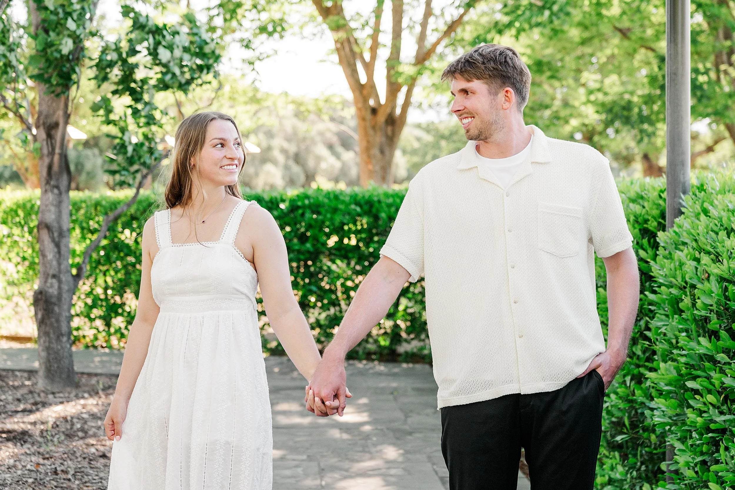 A smiling couple holds hands while walking on a sunny path surrounded by lush green trees and bushes. They are wearing casual, light-colored outfits.