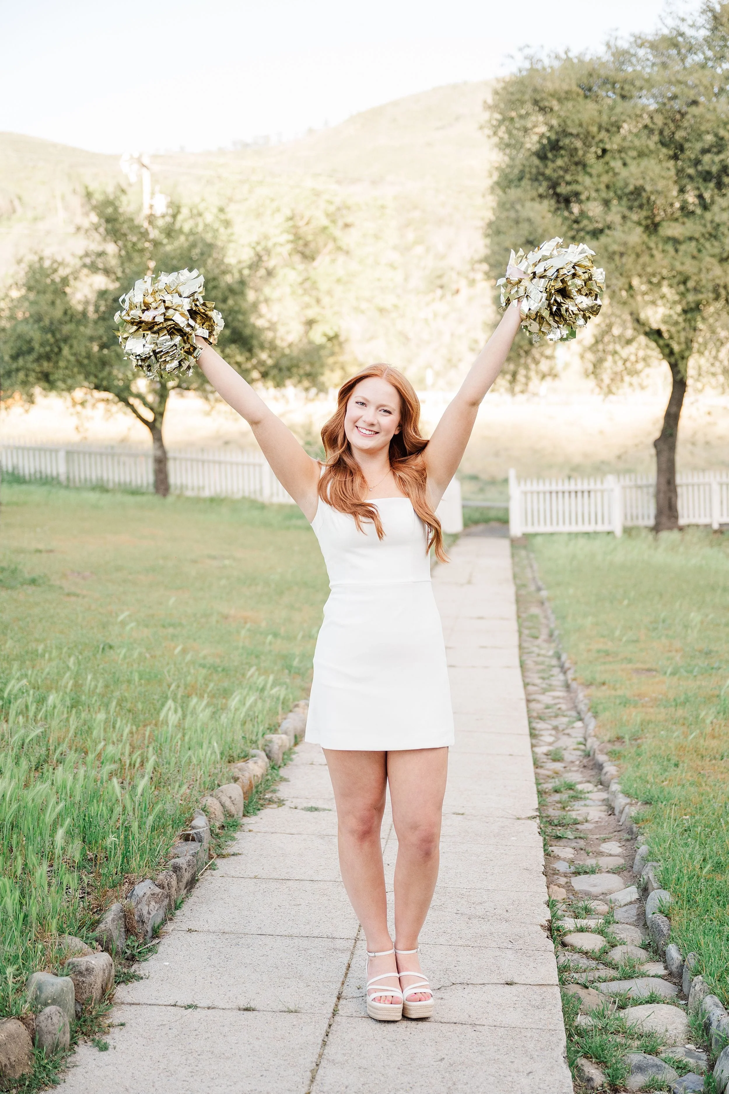 Young woman in a white dress joyfully raises pom-poms, standing on a paved path with green grass, trees, and a fence in a sunny landscape.