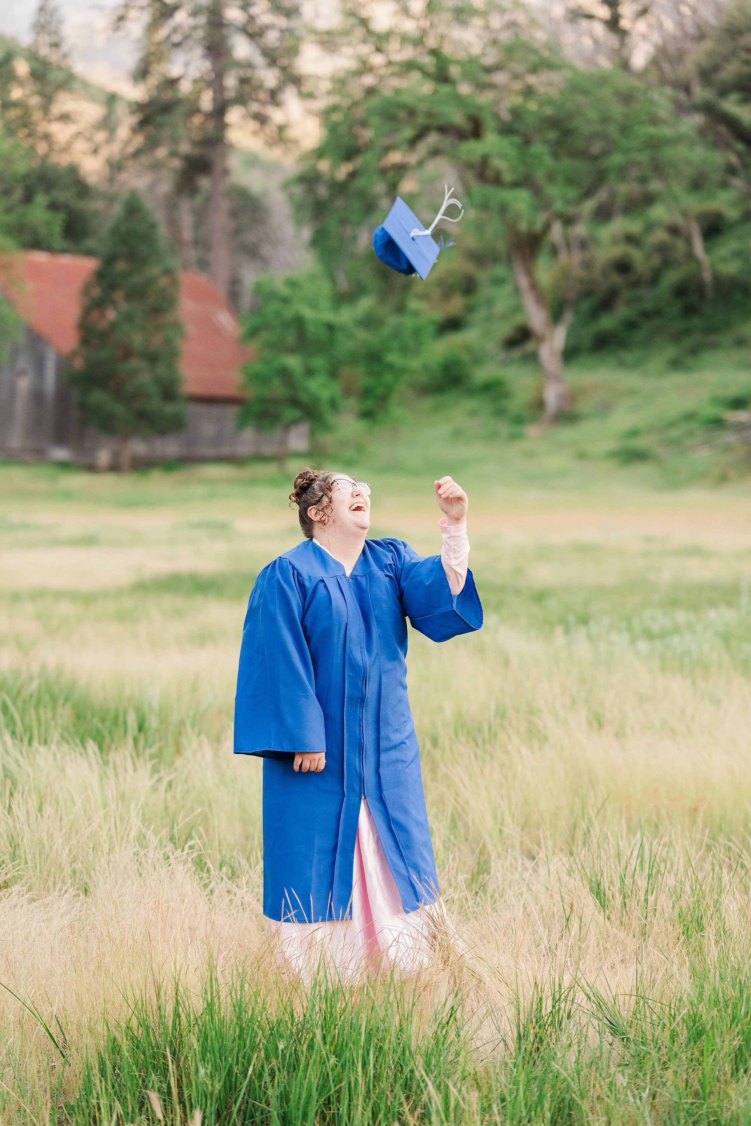 A graduate in a blue gown stands in a grassy field, joyfully tossing a cap in the air. A red-roofed barn and trees are in the background, evoking a sense of celebration and achievement.
