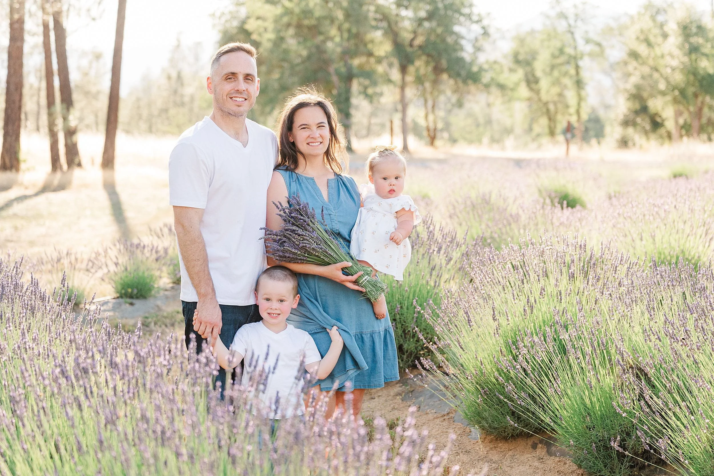 A smiling family of four stands in a lavender field under a sunny sky. The mother holds a baby and lavender bouquet, evoking warmth and happiness.