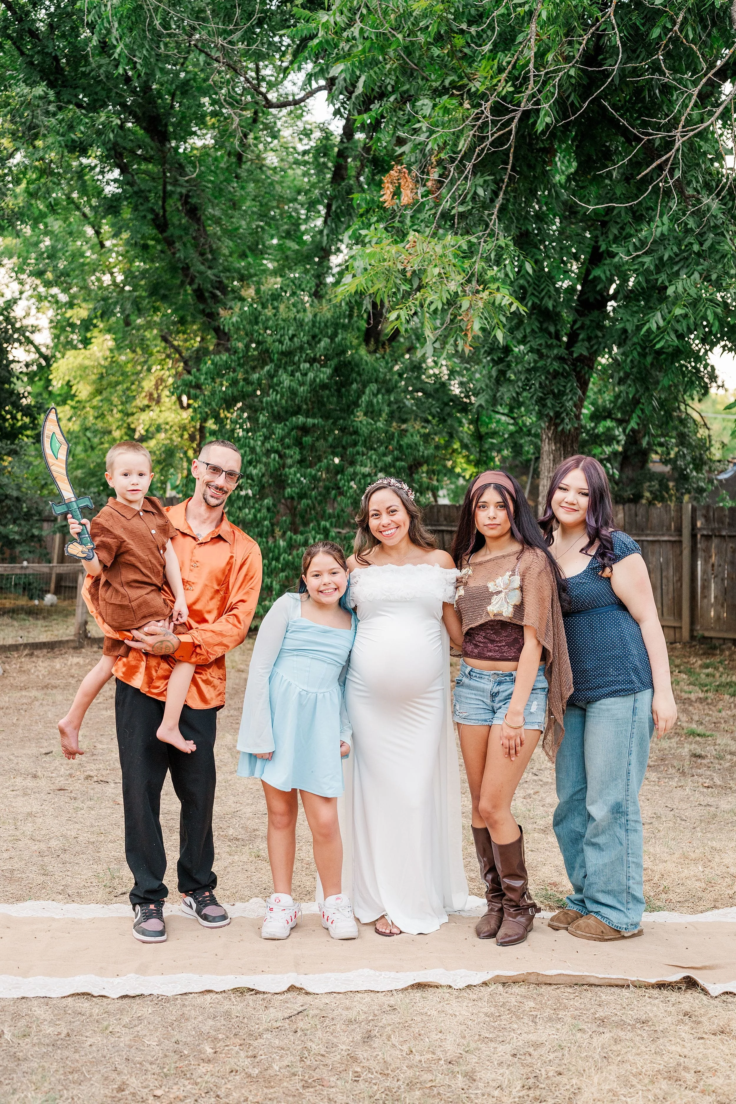 Outdoor family portrait: smiling parents, children, and a pregnant woman pose together in a leafy backyard, creating a warm, joyful atmosphere.
