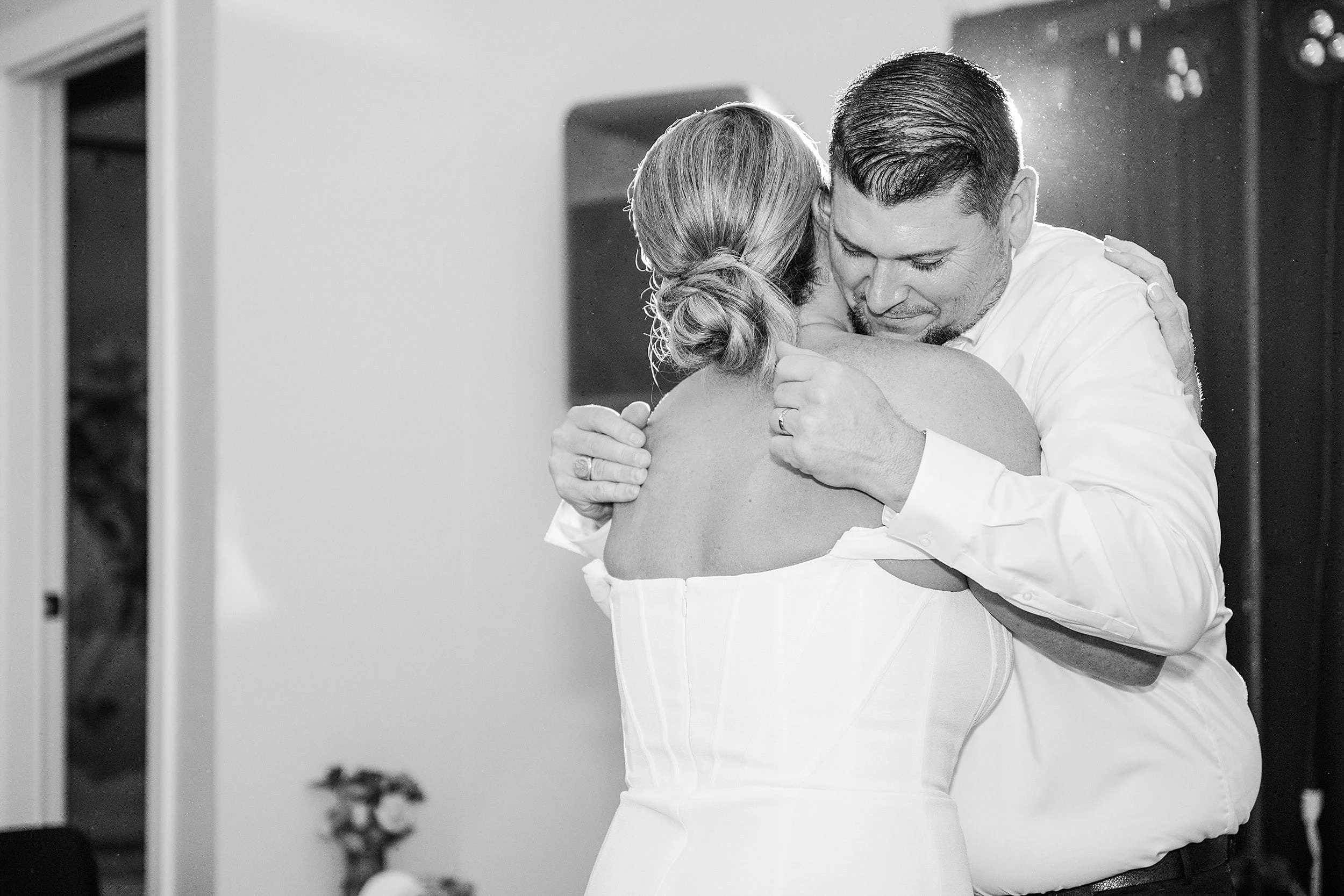A groom embraces his bride during their first dance, captured in black and white. The room is softly lit, creating an intimate and tender atmosphere.