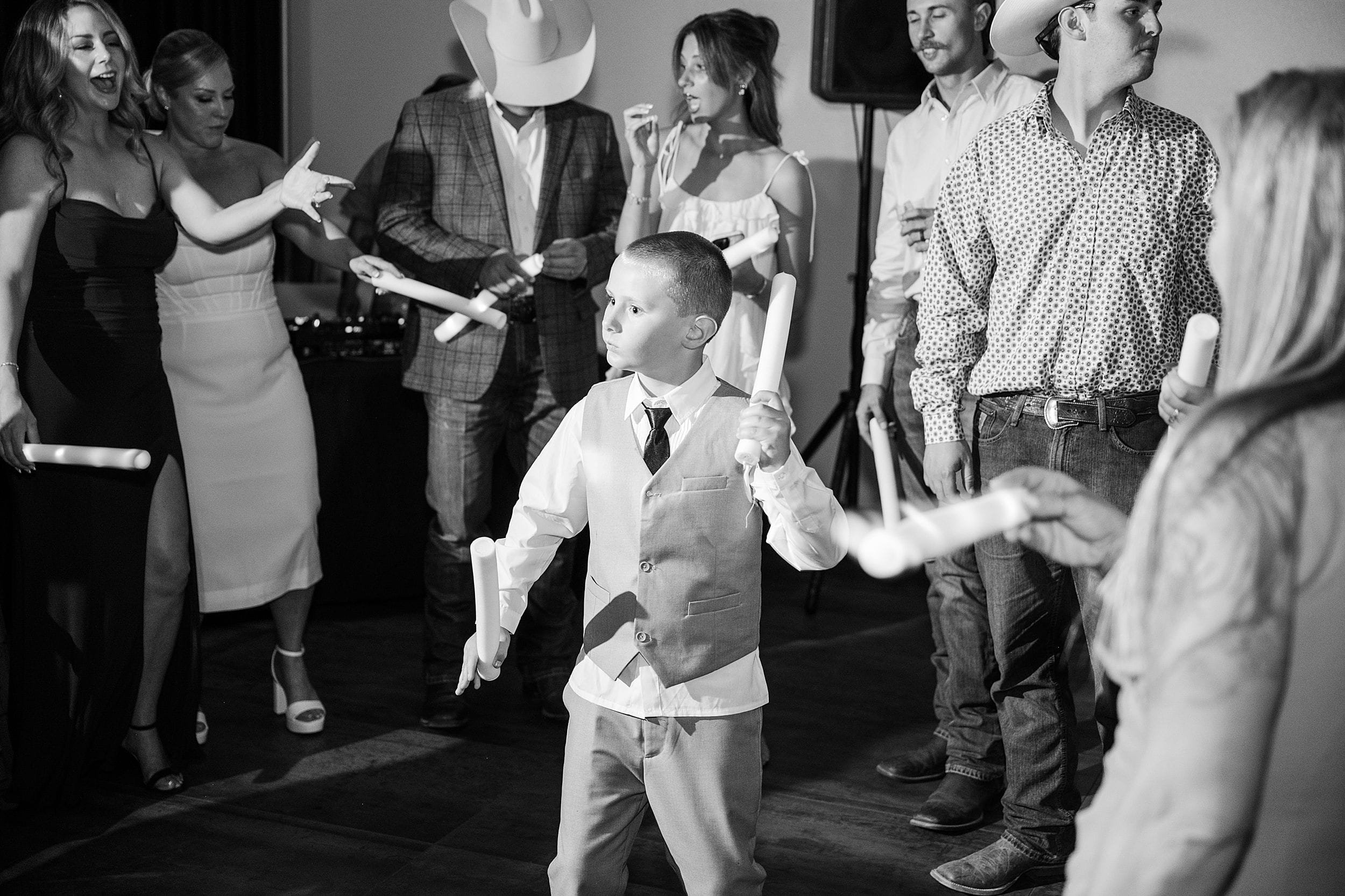 A young boy in a vest and tie holds glowing sticks, surrounded by people dancing joyfully at a party. The mood is lively and festive.