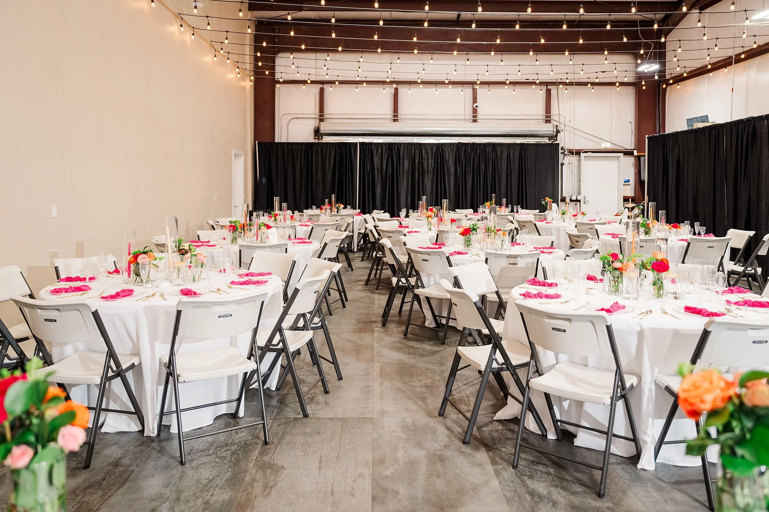 A large venue with round tables and white chairs set for an event. Tables have bright floral centerpieces and pink napkins. String lights create a festive atmosphere.