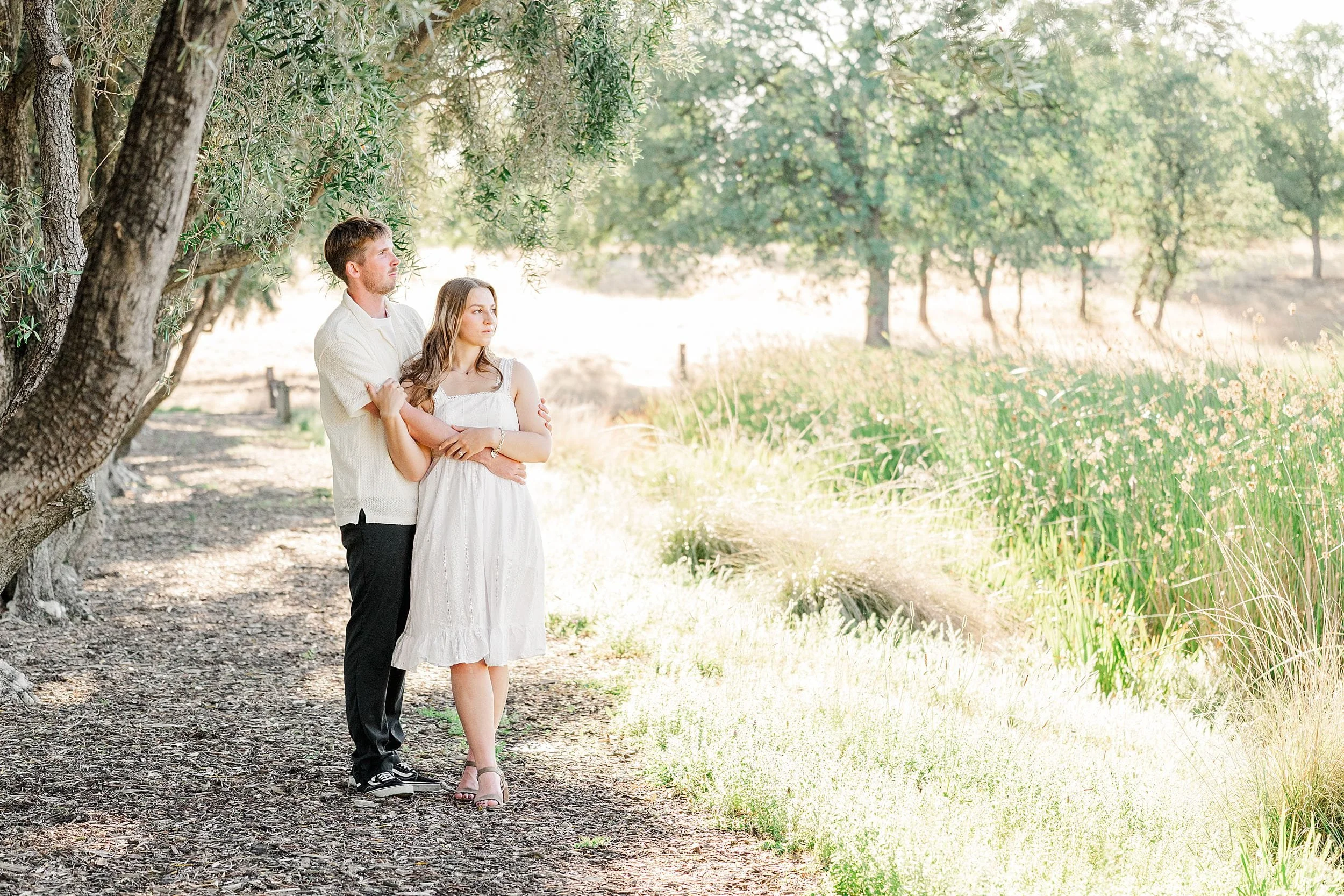 A couple stands on a sunlit path under trees, with the woman in a white dress and the man in a light shirt. They look serene, surrounded by greenery.