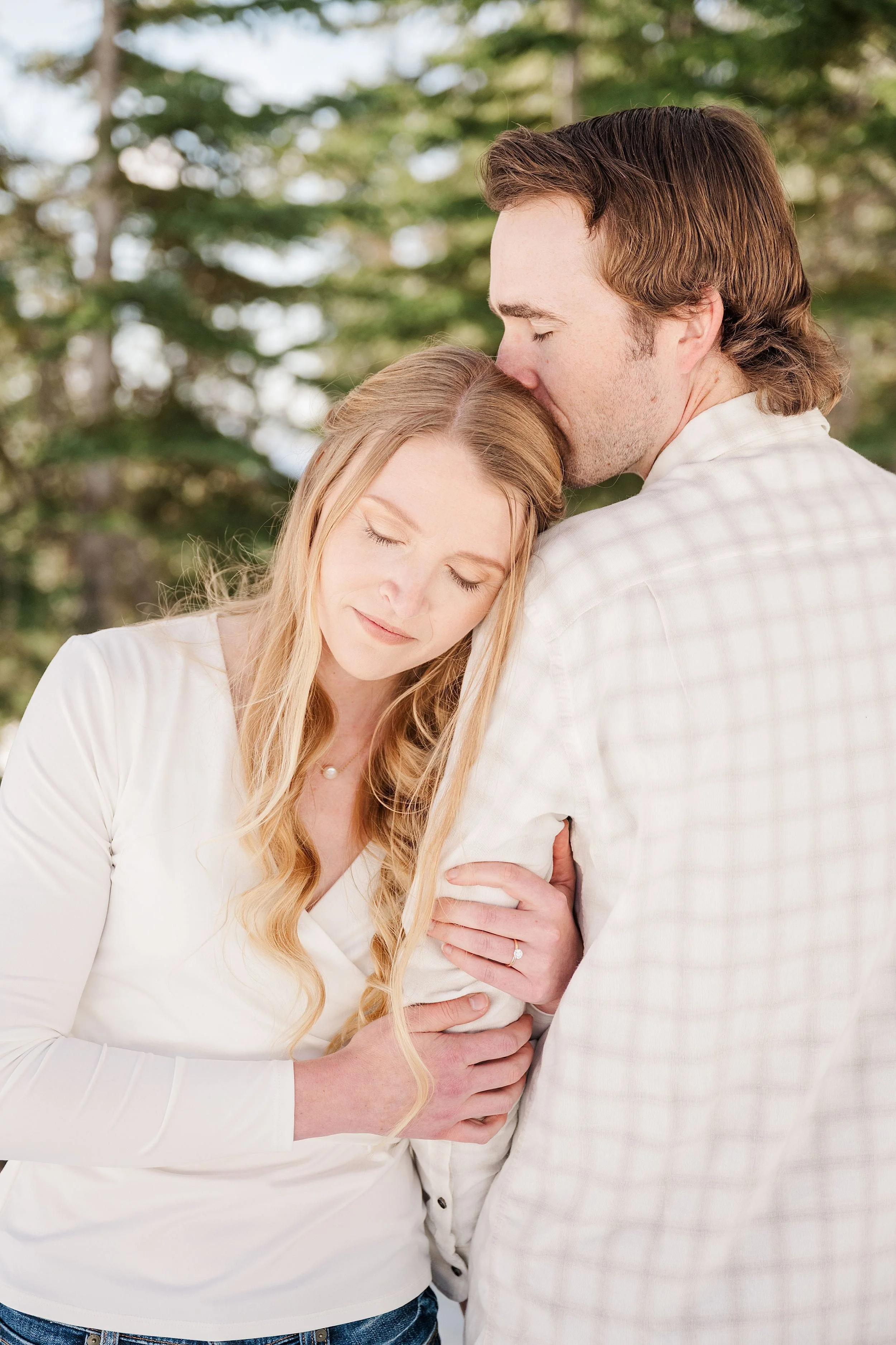 A couple embraces tenderly outdoors, with a woman resting her head on a man's shoulder. Both wear white, conveying warmth and intimacy amidst tall trees.