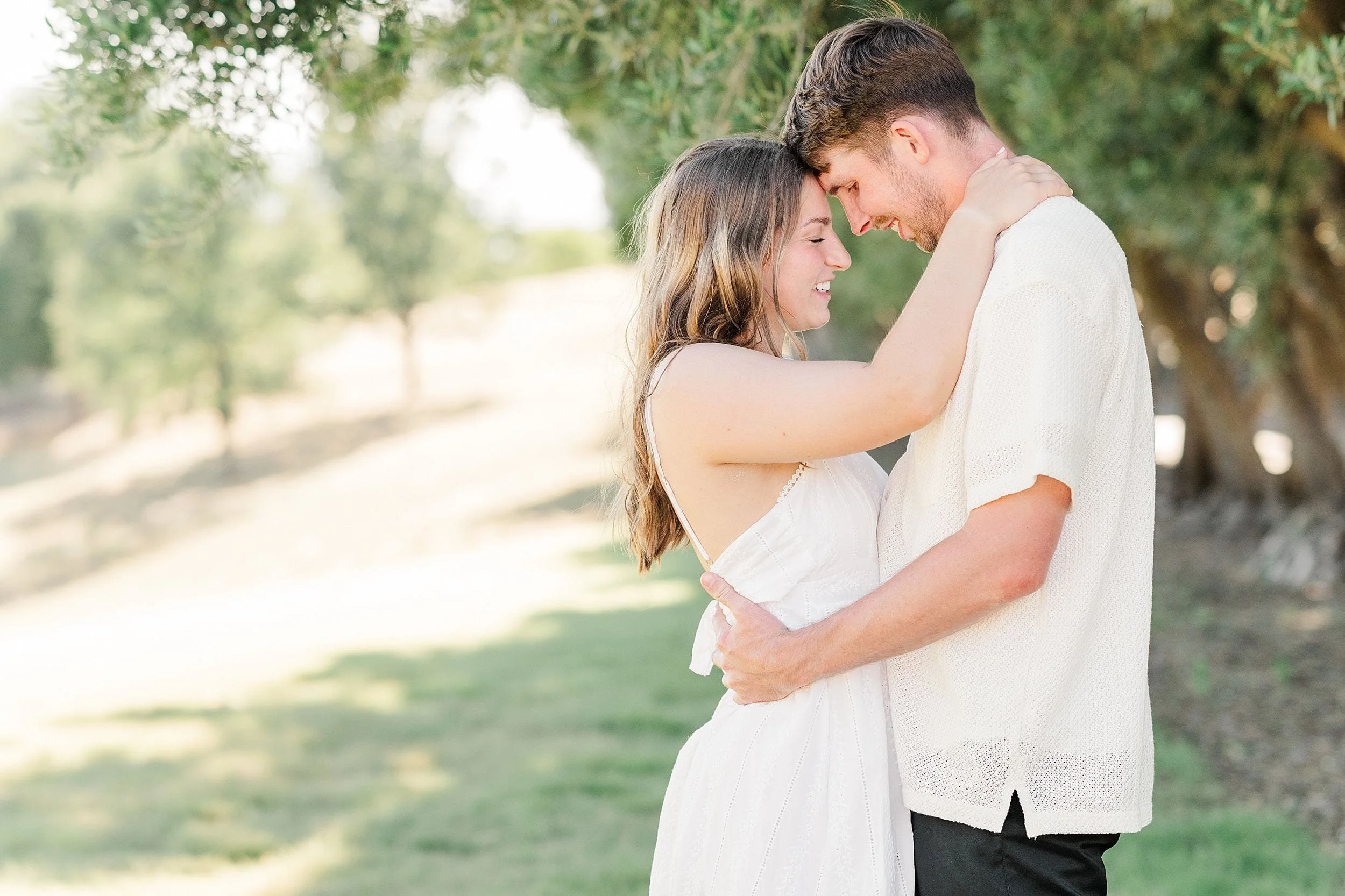 A couple embraces under a tree in a sunlit park. They gaze at each other with warm smiles, conveying intimacy and happiness. Both wear white attire.