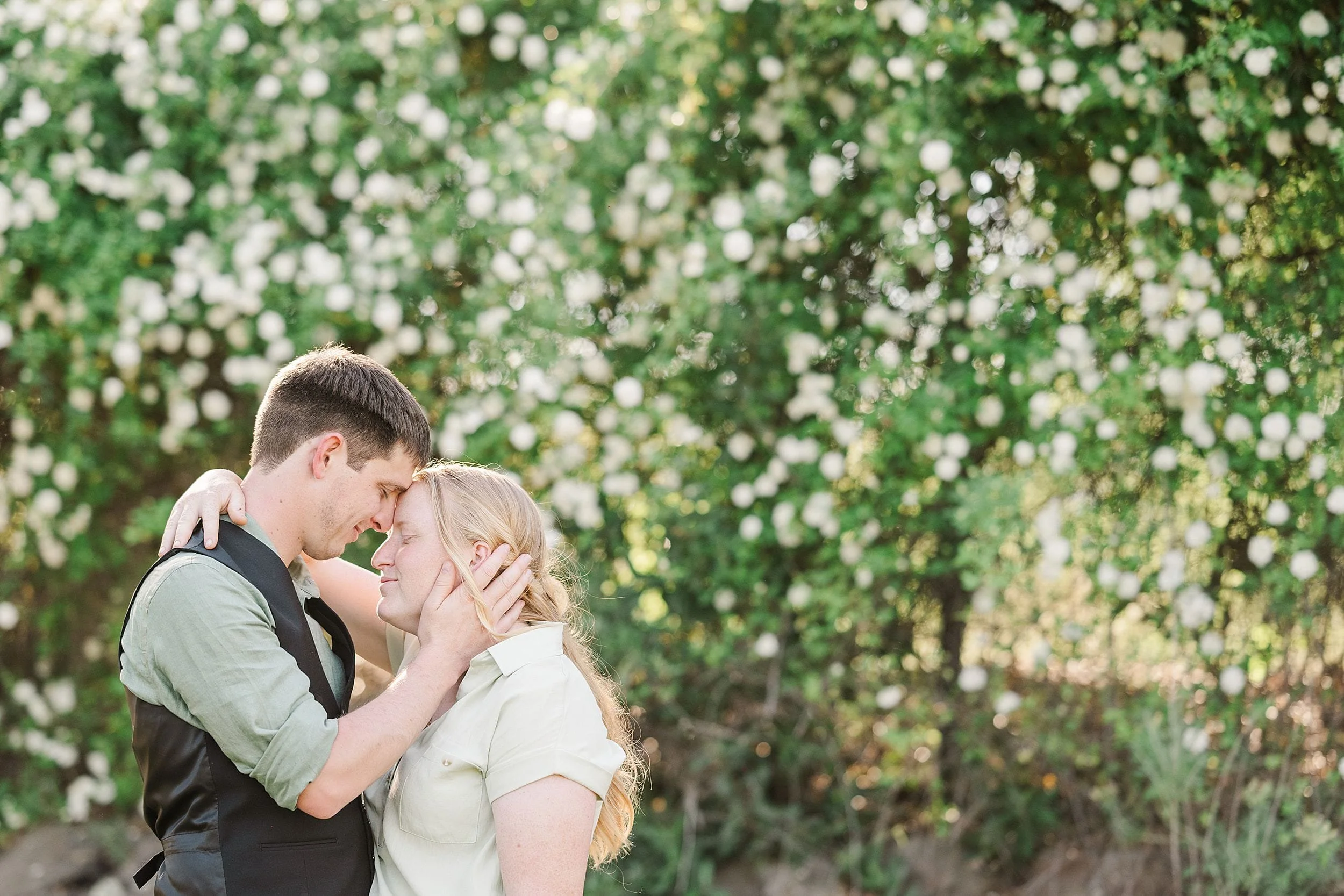 A couple embraces tenderly in front of a lush green backdrop adorned with white flowers, conveying love and serenity in a sunlit garden setting.