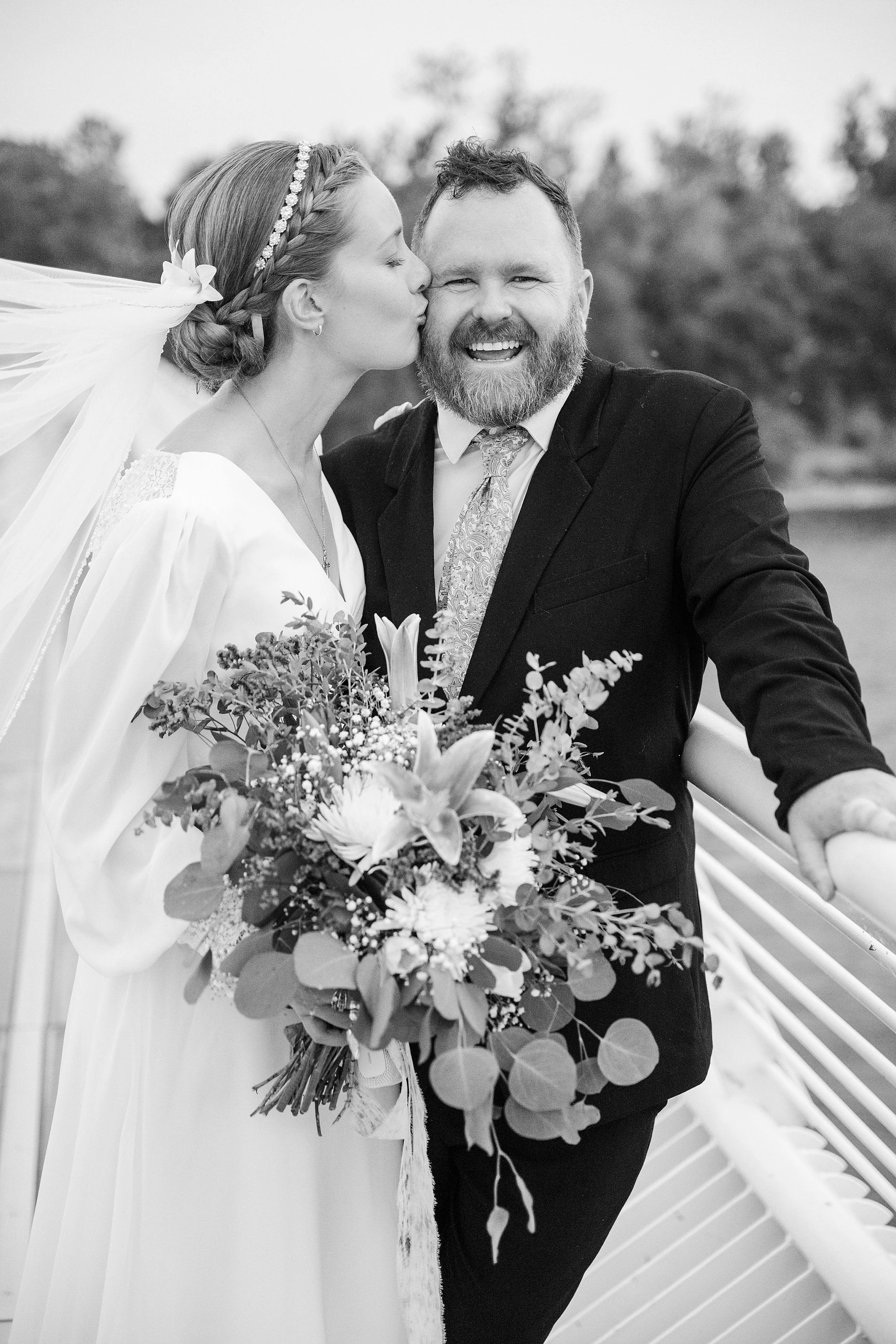 A joyful bride kisses a smiling groom on a bridge. She holds a large bouquet adorned with lilies and eucalyptus. The scene conveys romance and celebration.