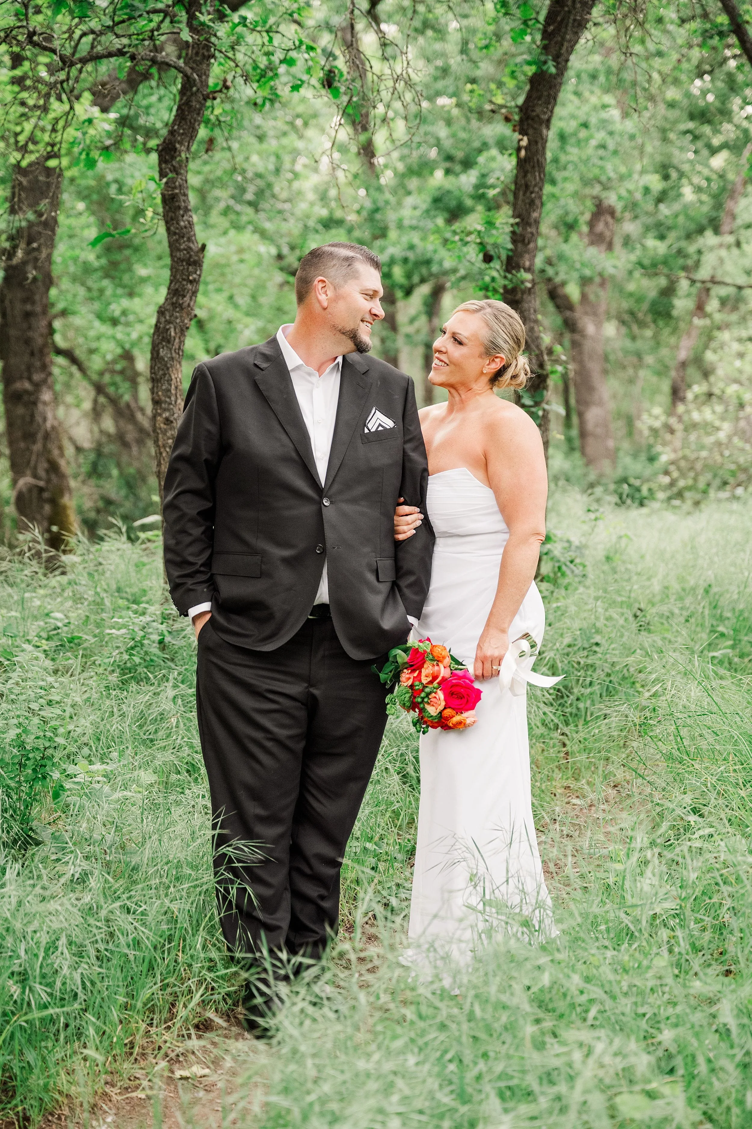 A happy couple walks arm-in-arm through a lush green forest. The bride, in a white dress, holds vibrant red flowers. The groom wears a black suit.
