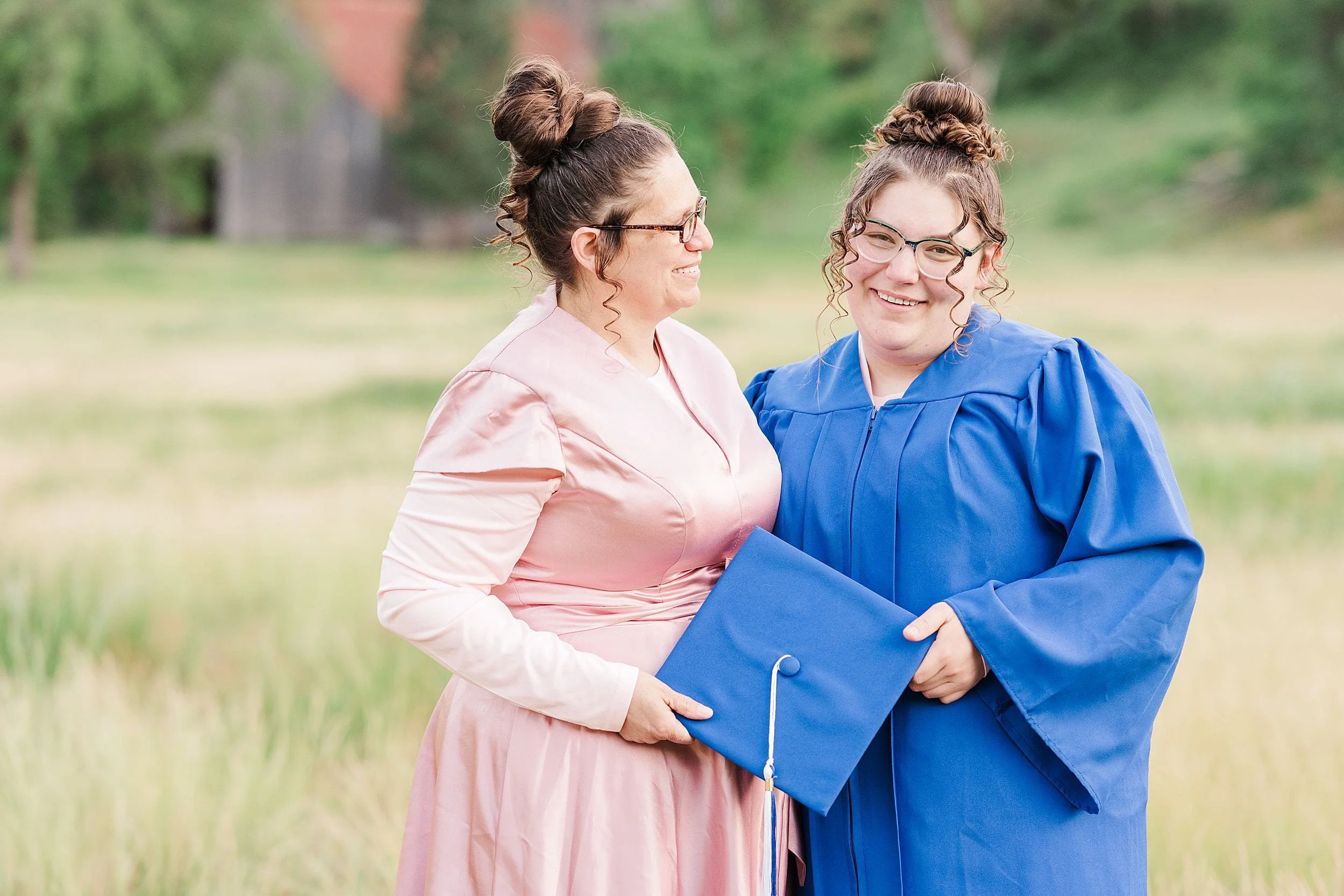 Two women stand in a grassy field, smiling warmly. One wears a pink dress, the other a blue graduation gown, holding a matching cap. Background is blurred greenery.