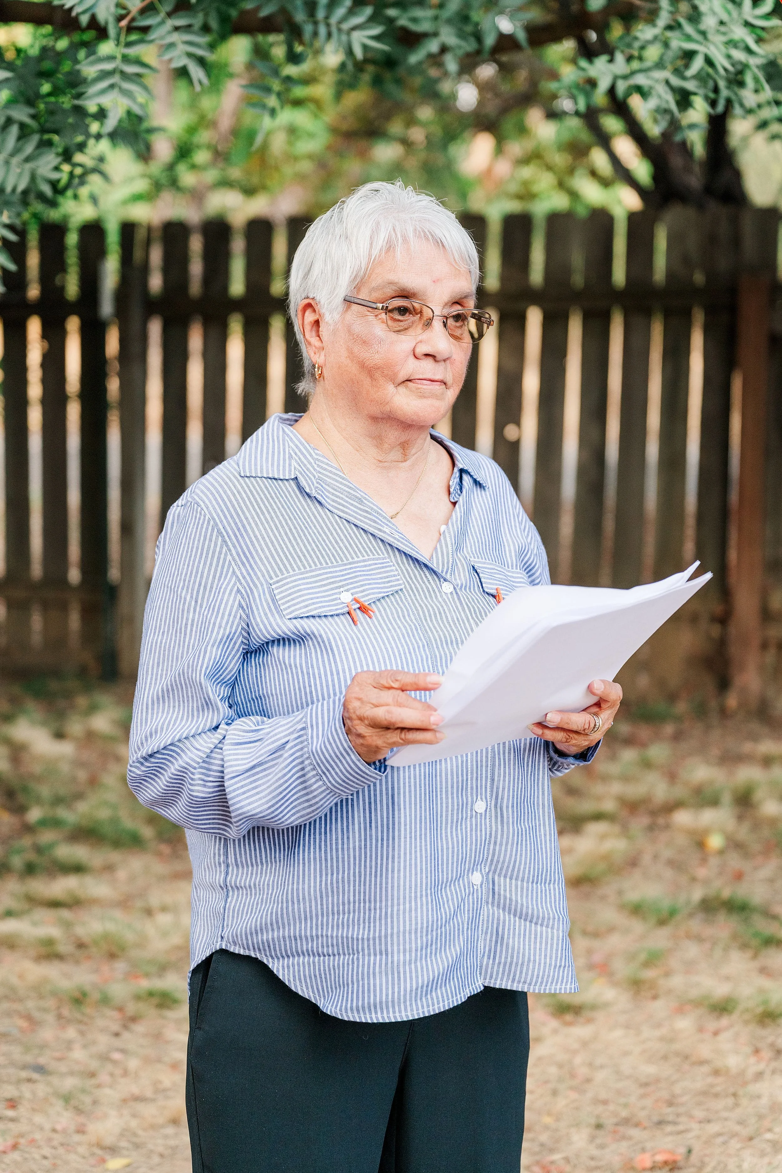 Older woman with short white hair and glasses reads papers outdoors before a wooden fence and leafy garden, conveying a calm, focused mood.
