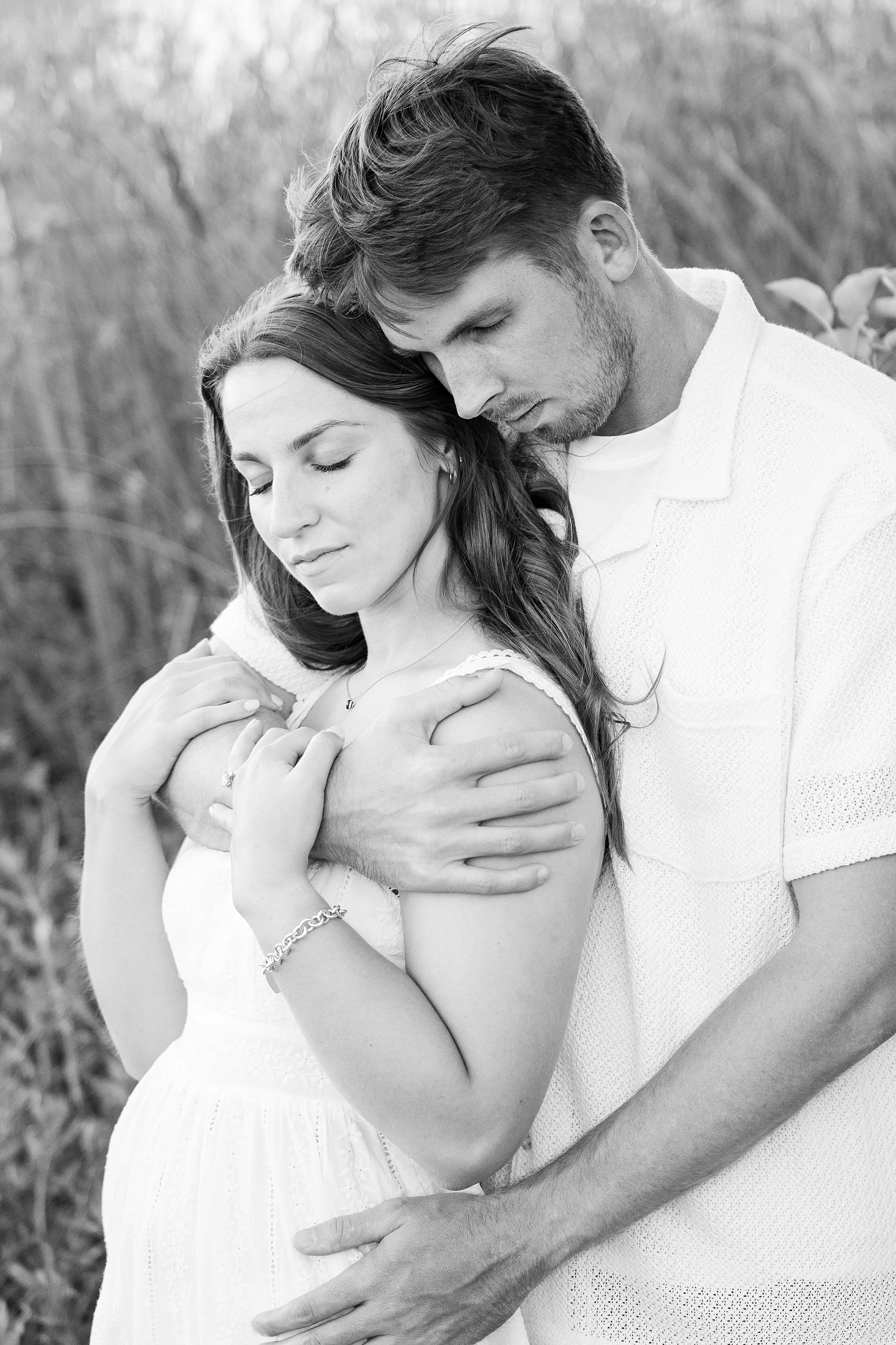 A couple tenderly embraces outdoors. The woman in a white dress closes her eyes, leaning into the man, who wraps his arms around her, exuding warmth.