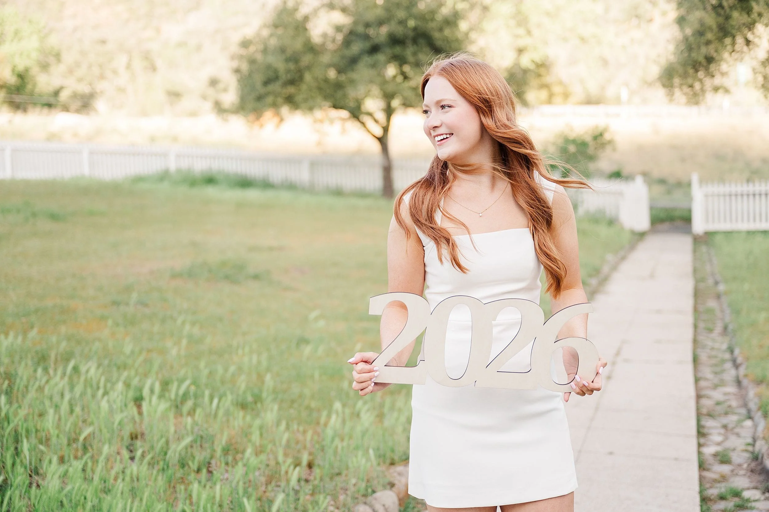 A young woman in a white dress stands smiling in a grassy yard, holding a "2026" sign. The background features a tree and white fence, conveying a joyful, celebratory tone.
