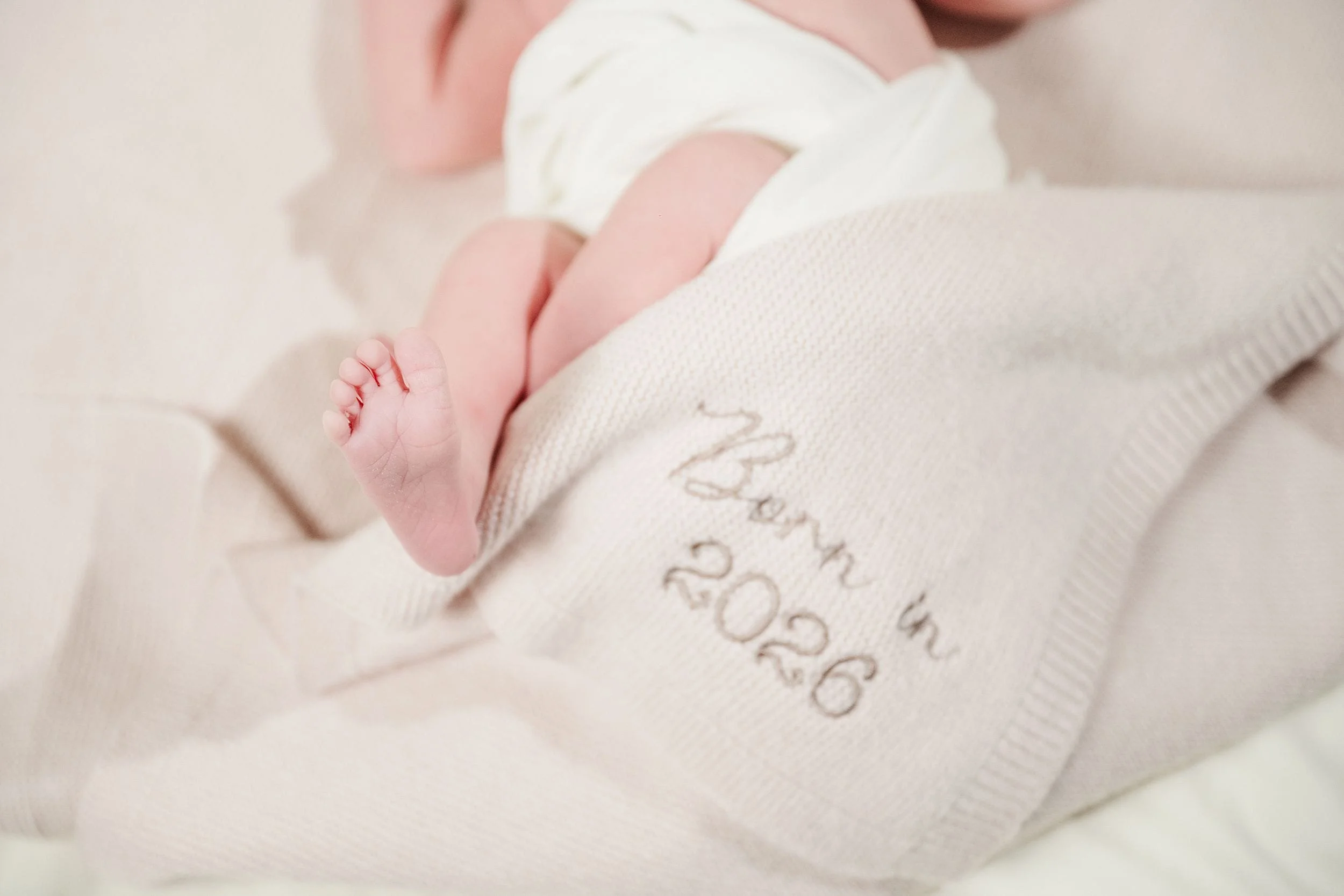 Newborn wrapped in a soft beige blanket embroidered with "Born in 2026." Baby's small feet peek out, conveying warmth and innocence.