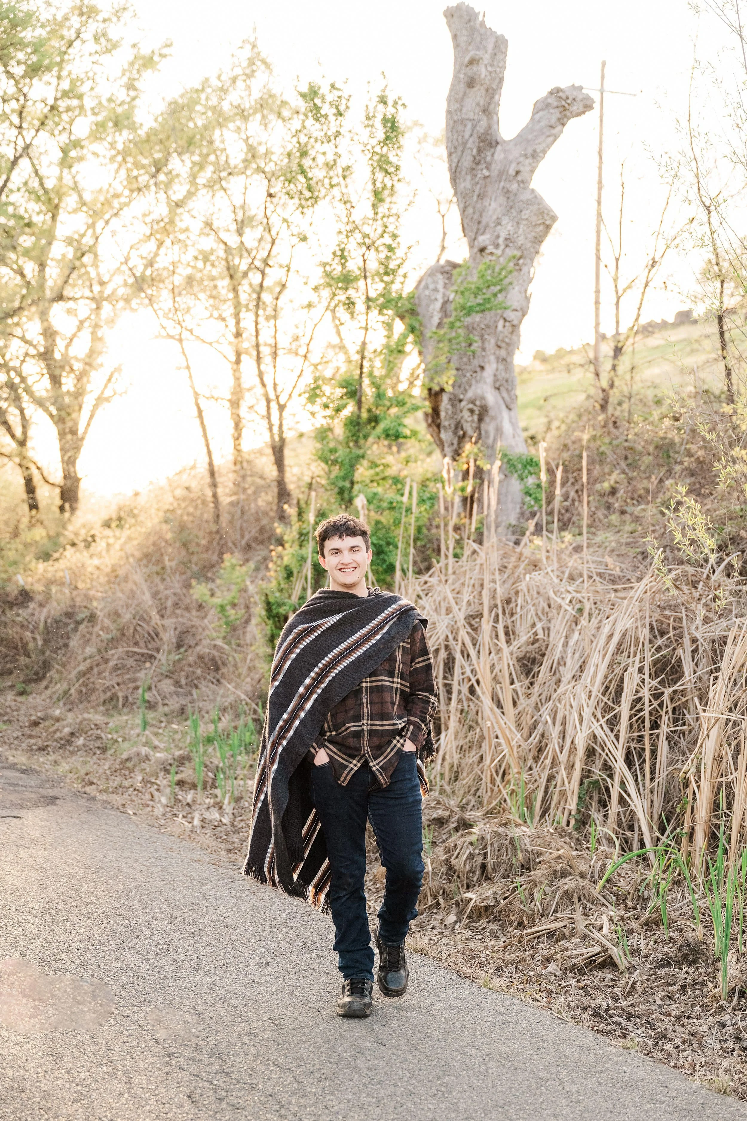 Young man walking outdoors on a paved trail, smiling, wearing a plaid shirt, dark jeans, hiking boots, and a striped shawl, with dry plants, trees, and a large weathered tree trunk in the background during sunset.