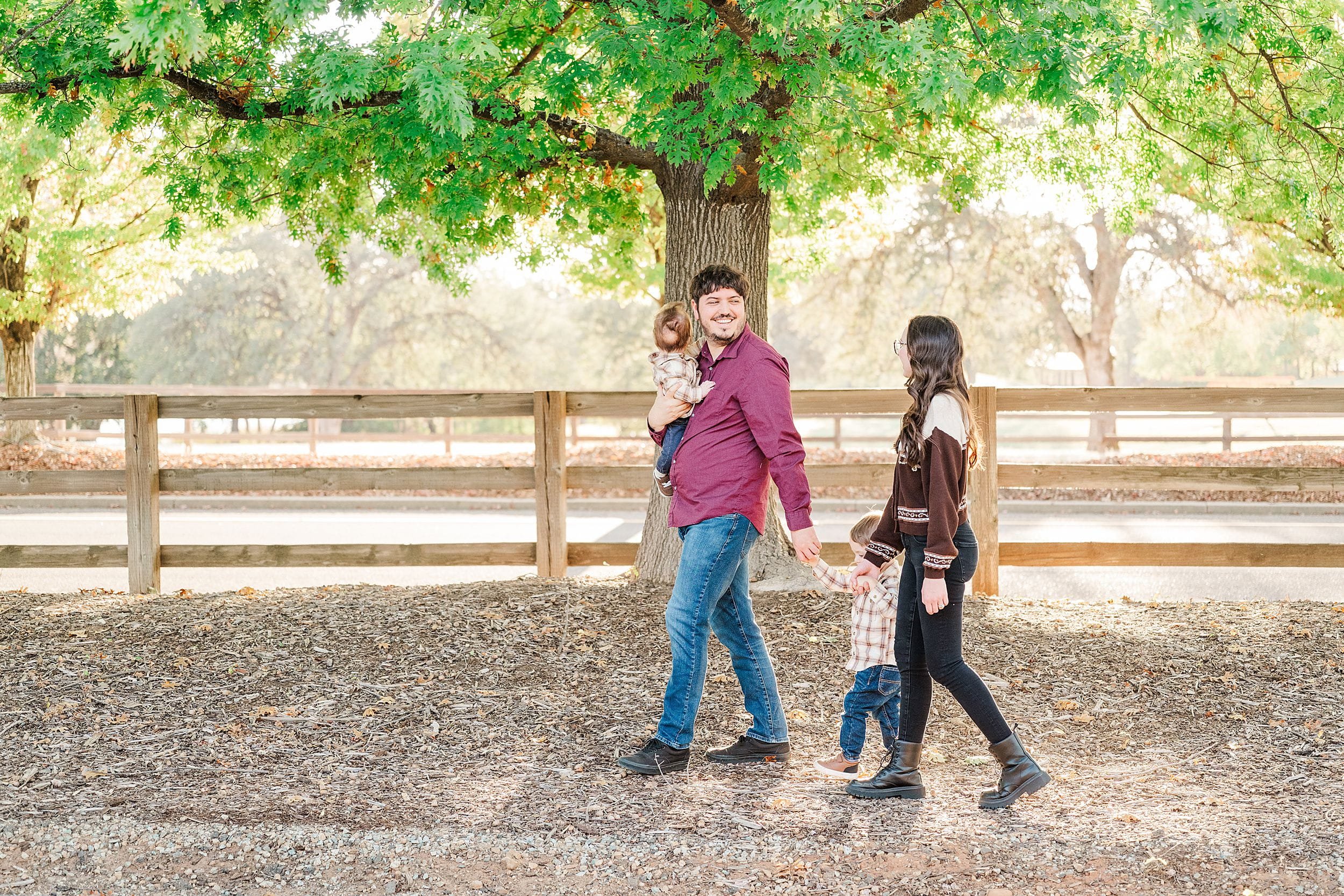 A smiling father carries a young child as he walks with a woman and another child beneath a leafy tree by a wooden fence, in warm autumn light.