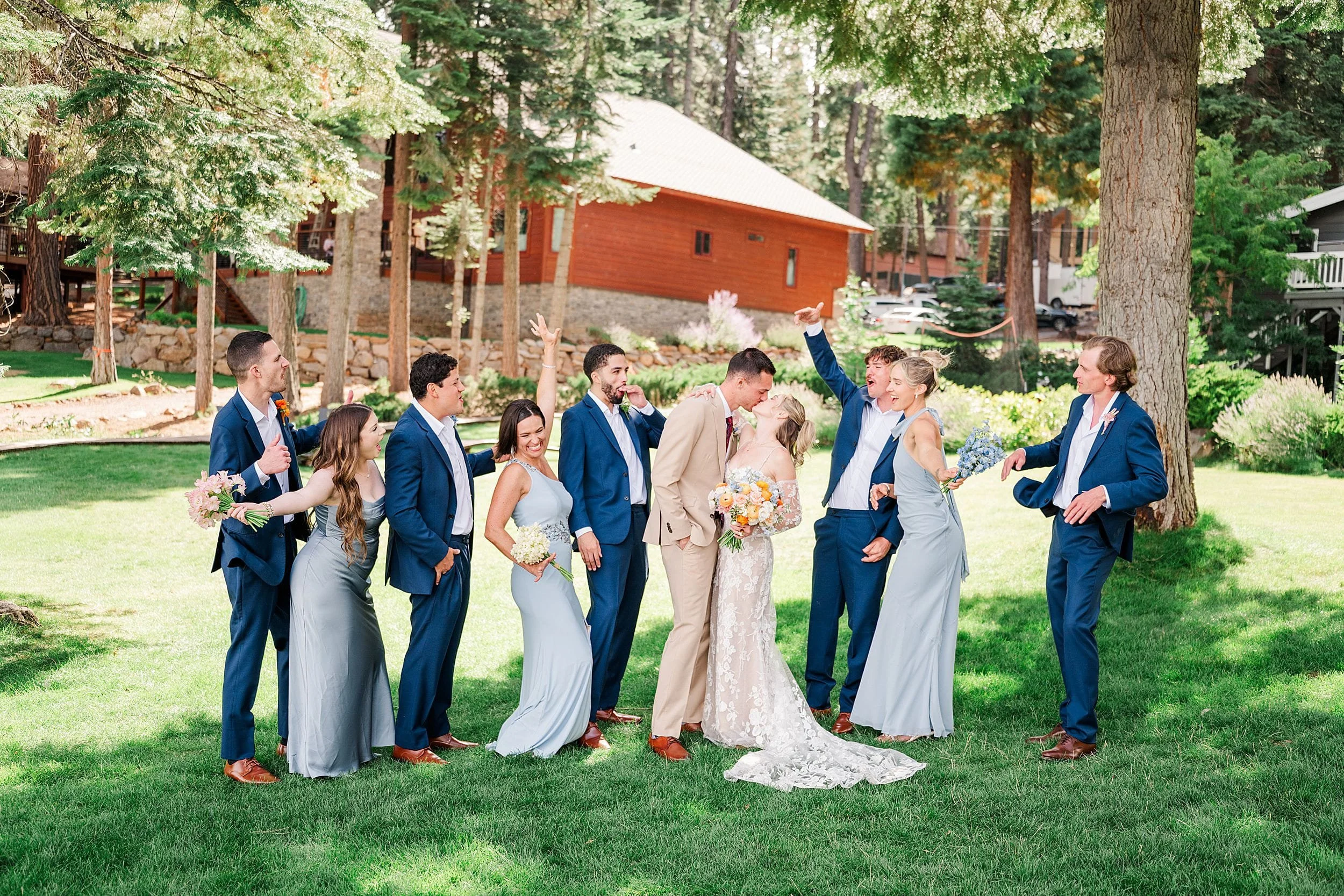Outdoor wedding party poses on a sunny lawn near a red cabin, with bride and groom surrounded by smiling bridesmaids and groomsmen.