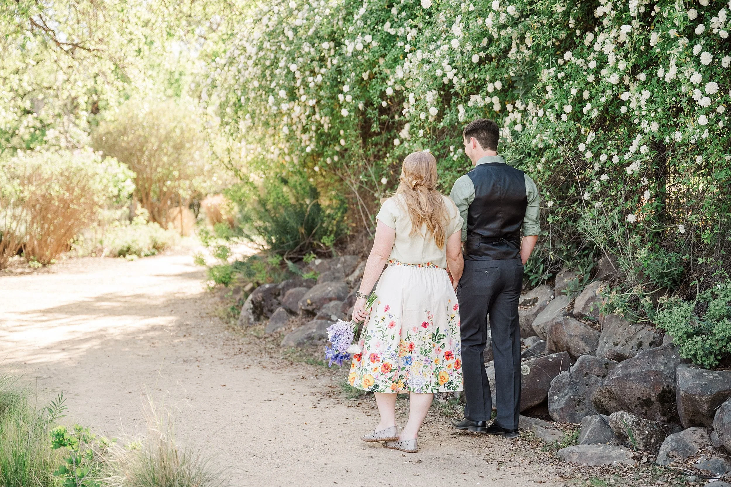 A couple walks hand in hand along a sunlit garden path lined with lush greenery and blooming white flowers. The scene conveys tranquility and romance.