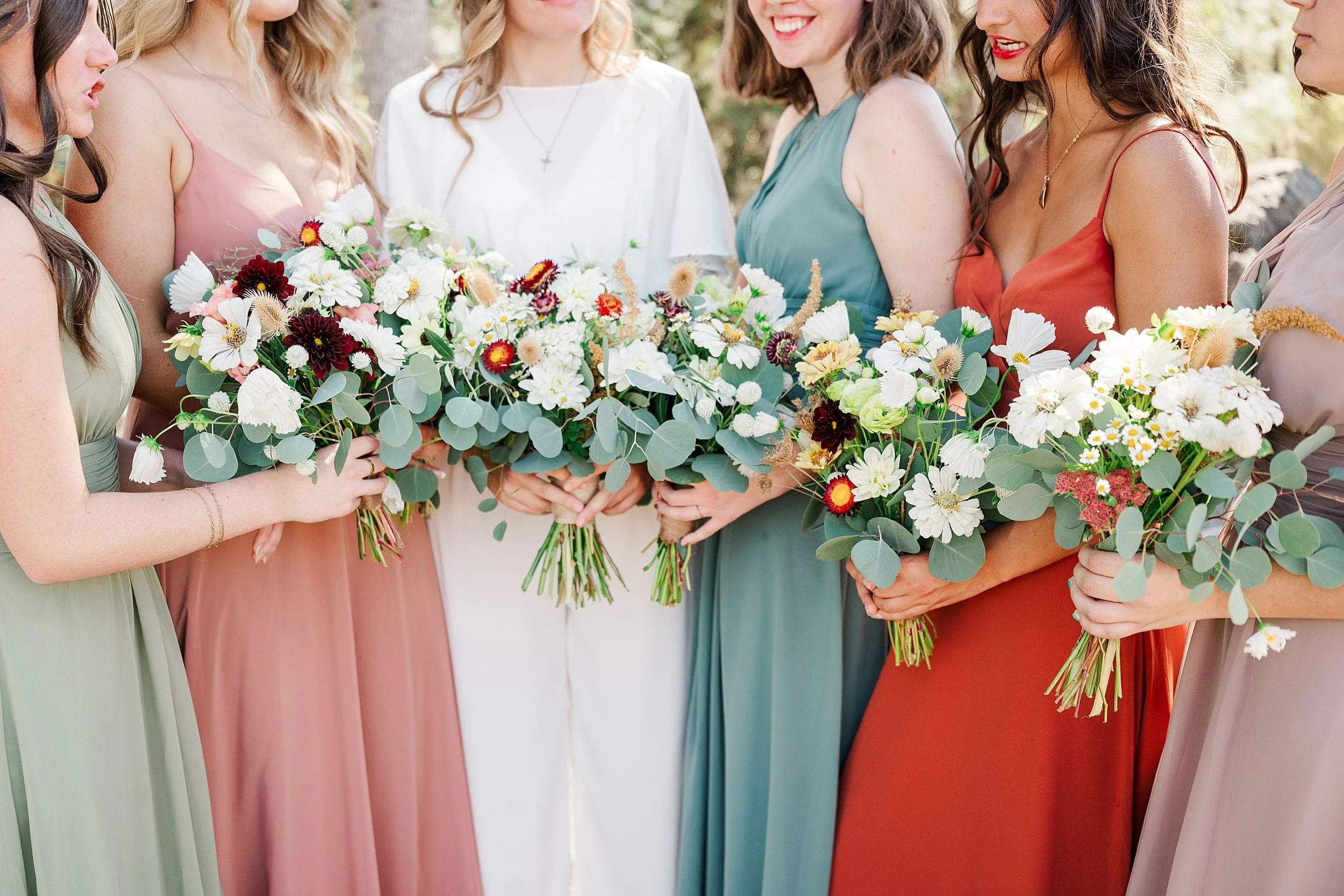 Bridesmaids in pastel and jewel-toned dresses hold lush bouquets of white flowers, roses, and greenery at an outdoor wedding, creating a joyful, elegant scene.