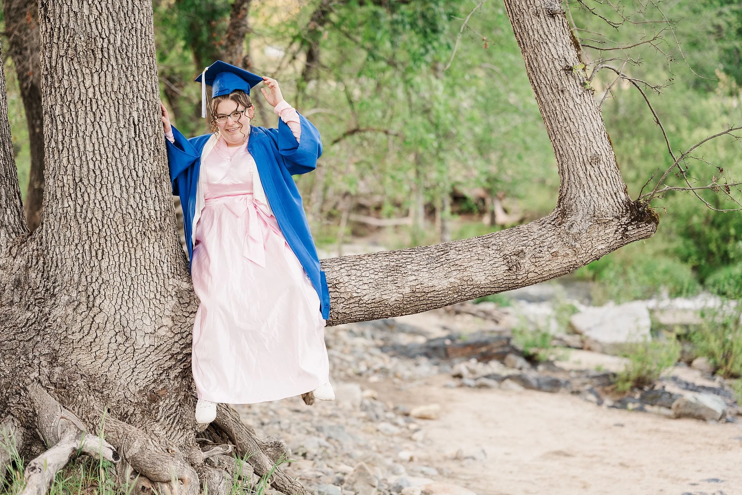 A person in a blue graduation gown and cap sits playfully on a tree branch, surrounded by greenery. They smile, exuding a joyful, celebratory mood.