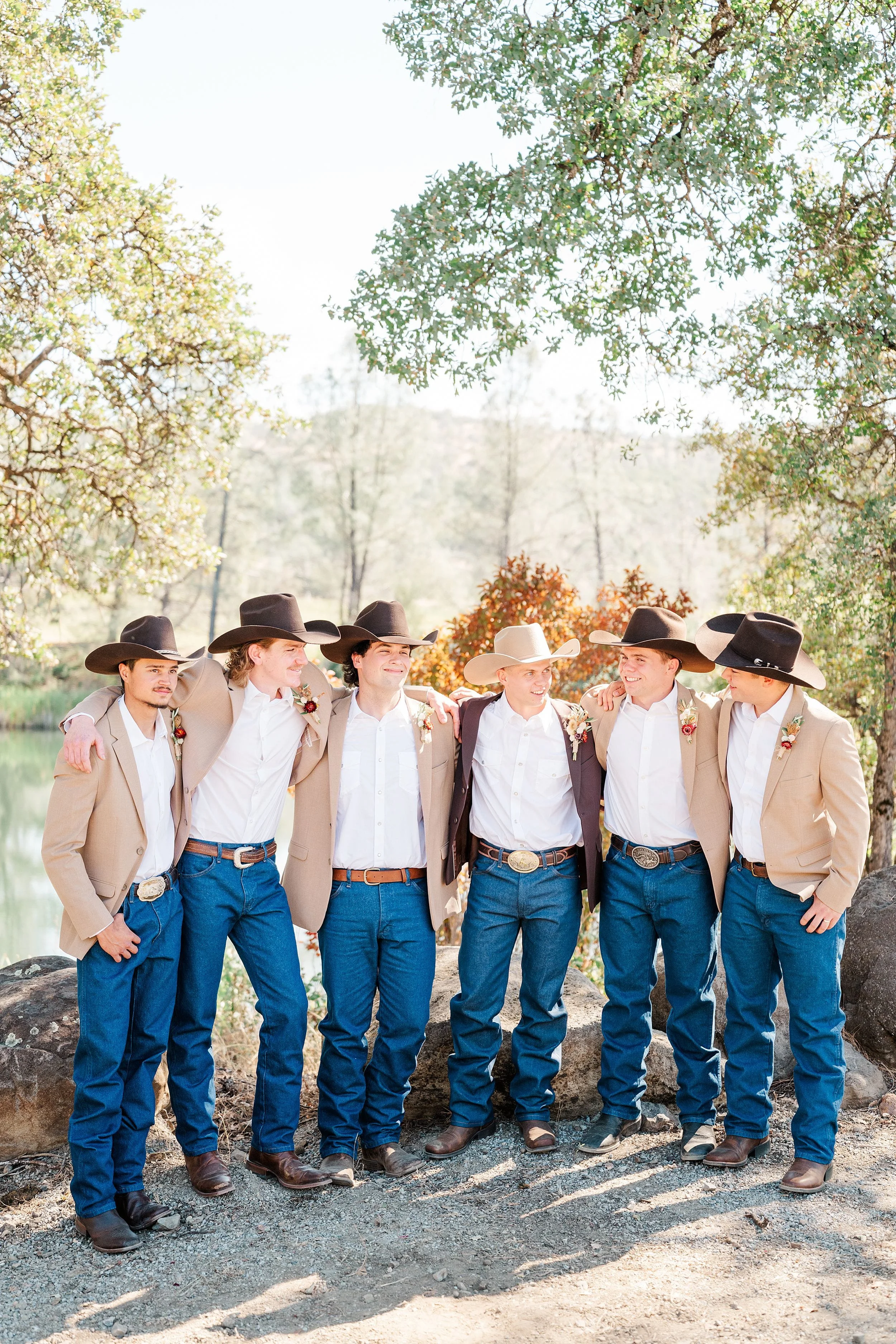 Six young men in cowboy hats, boots, jeans, and jackets stand arm in arm beneath leafy trees by a lake, smiling in warm, rustic outdoor light.
