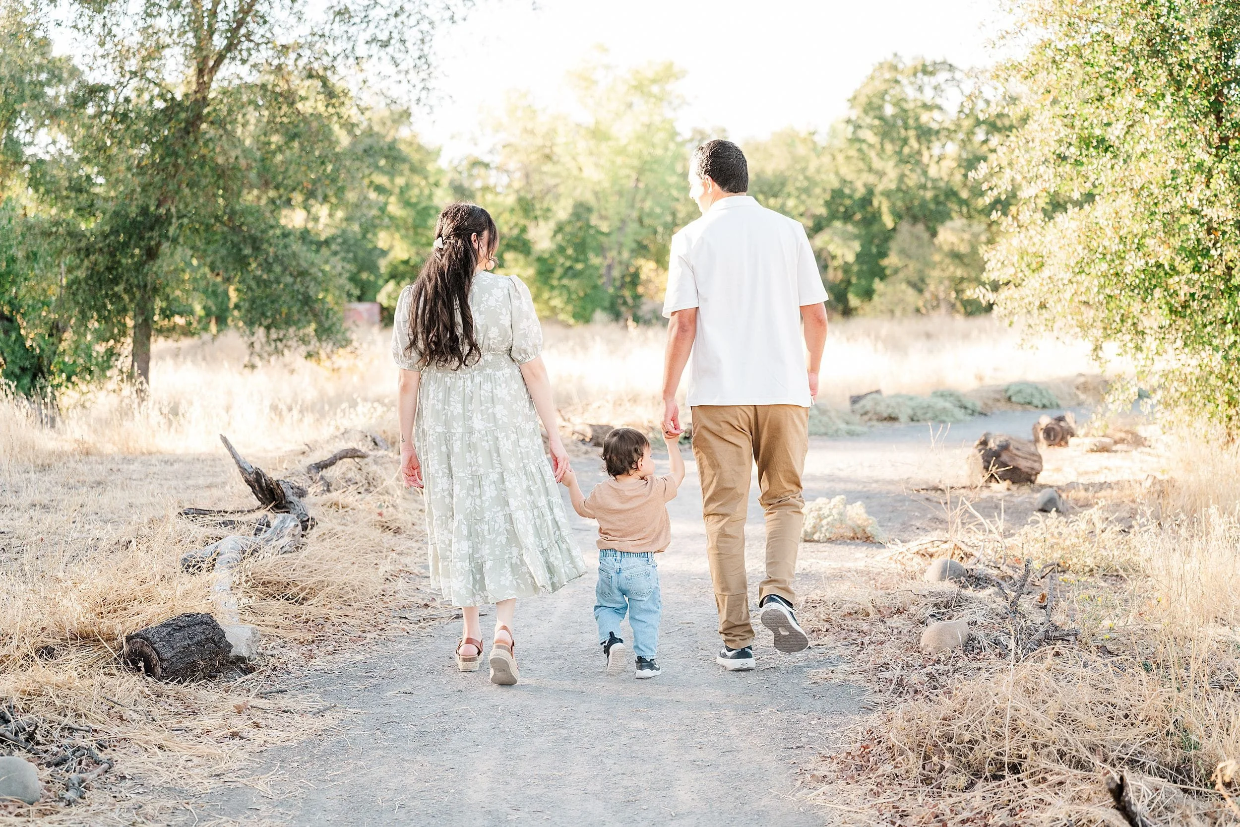 A mother and father hold their toddler’s hands as they walk a sunlit woodland path, surrounded by dry grass and trees in a peaceful family moment.