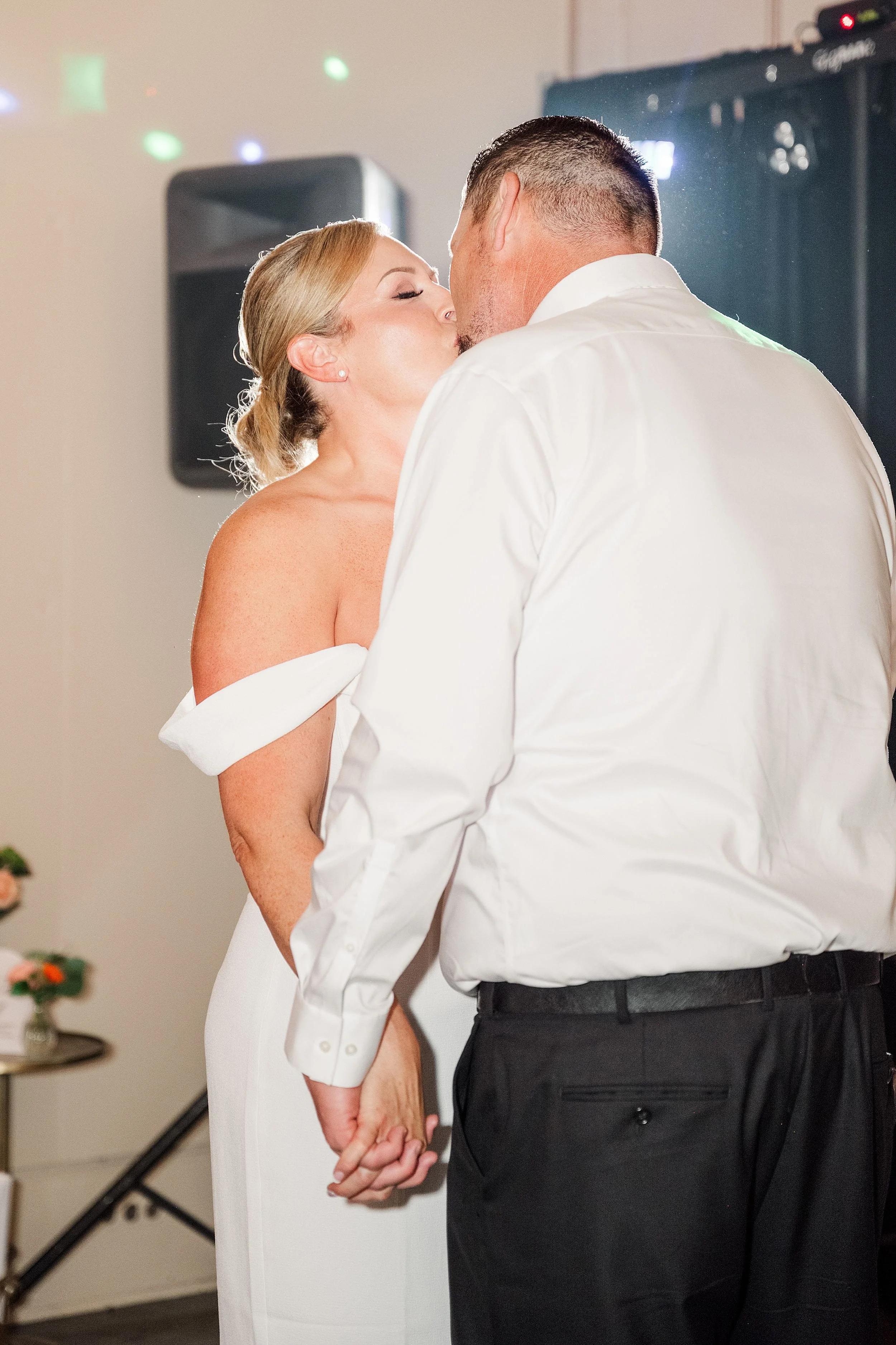 A couple shares a tender kiss at a wedding reception. The woman in a white dress holds hands with the man in a white shirt and black pants.