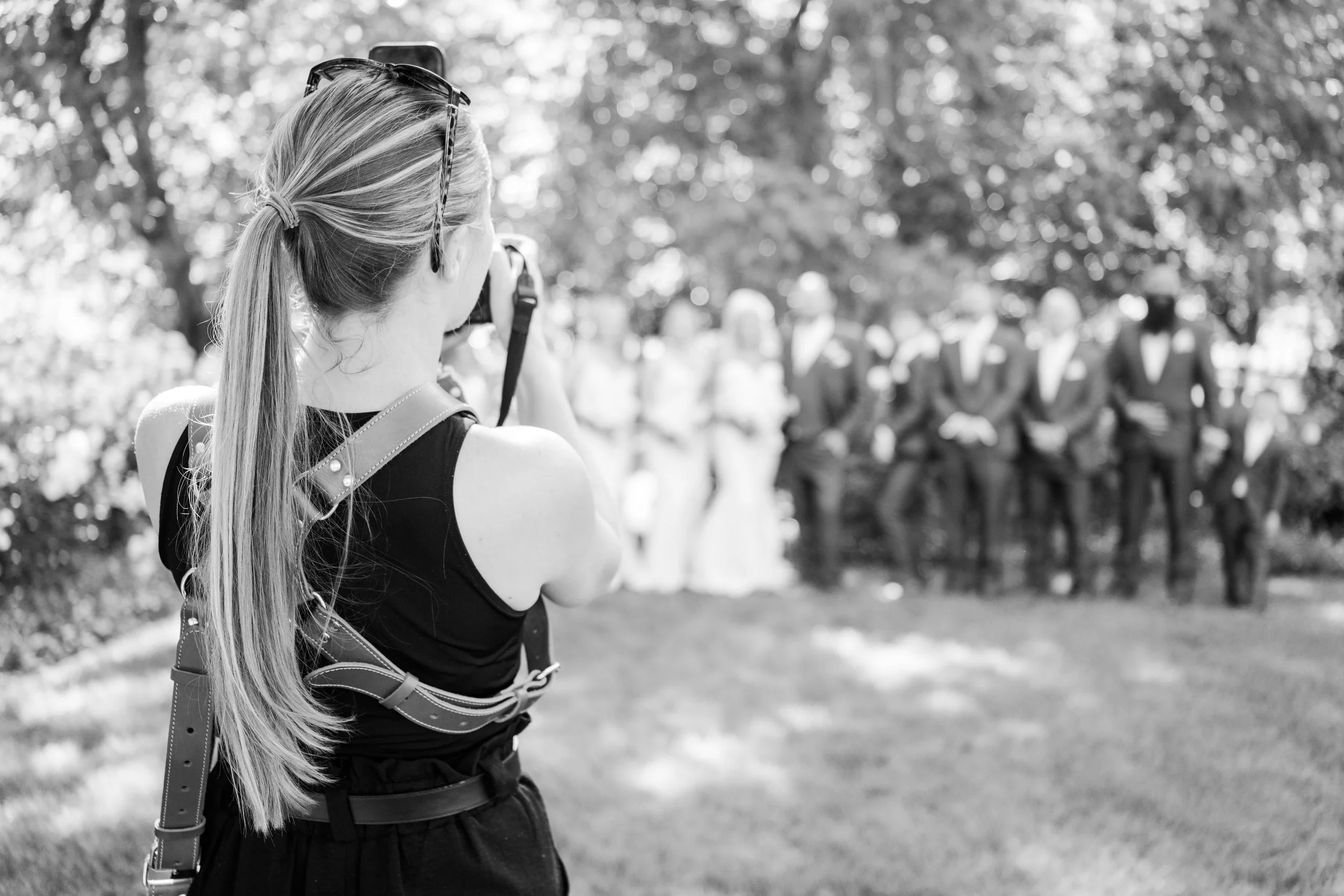 A woman with long hair tied in a ponytail, taking a photo with a camera at The Barn at Pheasant Trail. She is wearing sunglasses on her head and a black sleeveless top. The background shows a wedding party gathered with trees and sunlight.