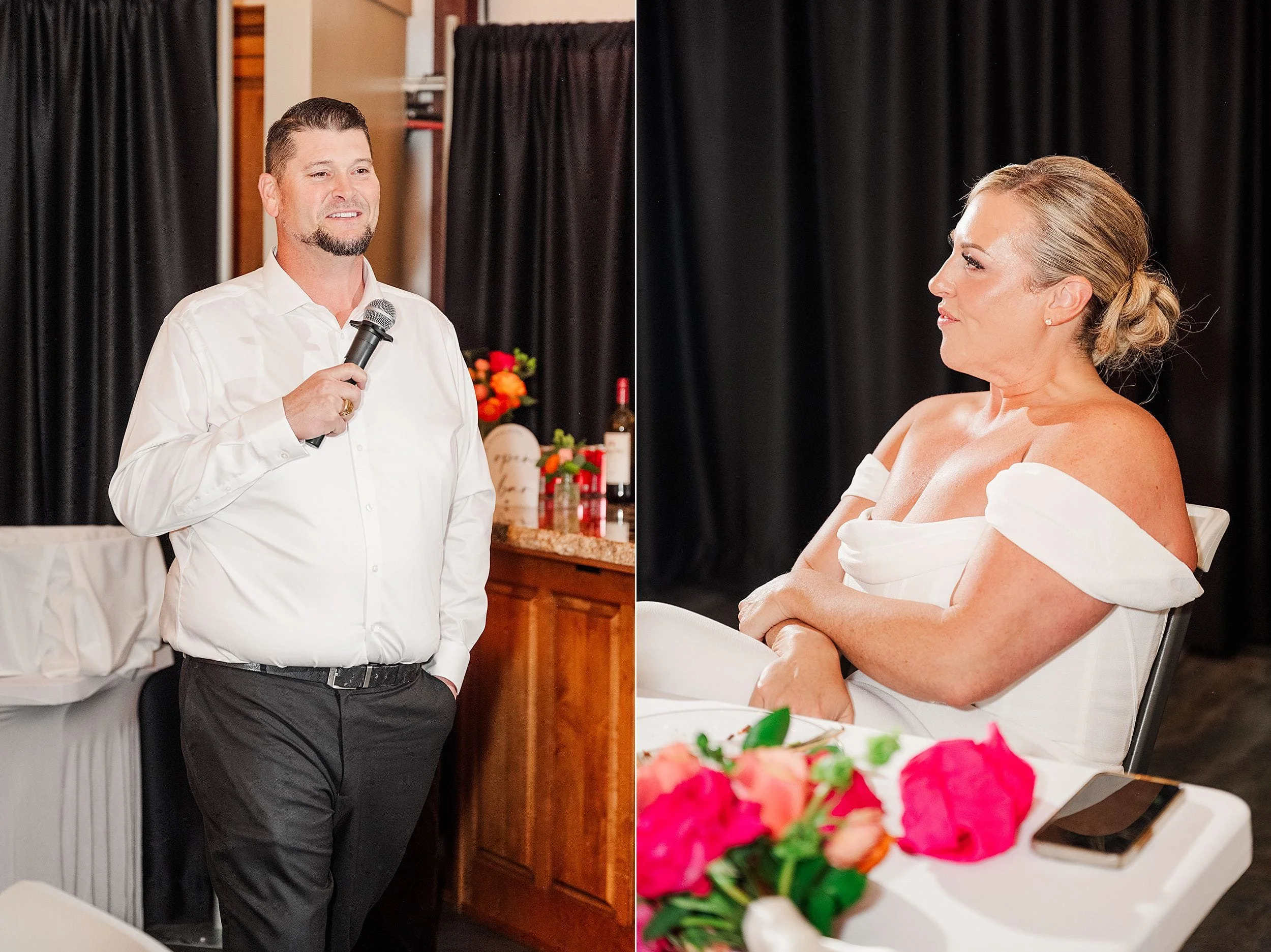 A man in a white shirt speaks into a microphone, smiling, while a woman in an off-shoulder dress listens attentively, seated at a table with vibrant flowers.
