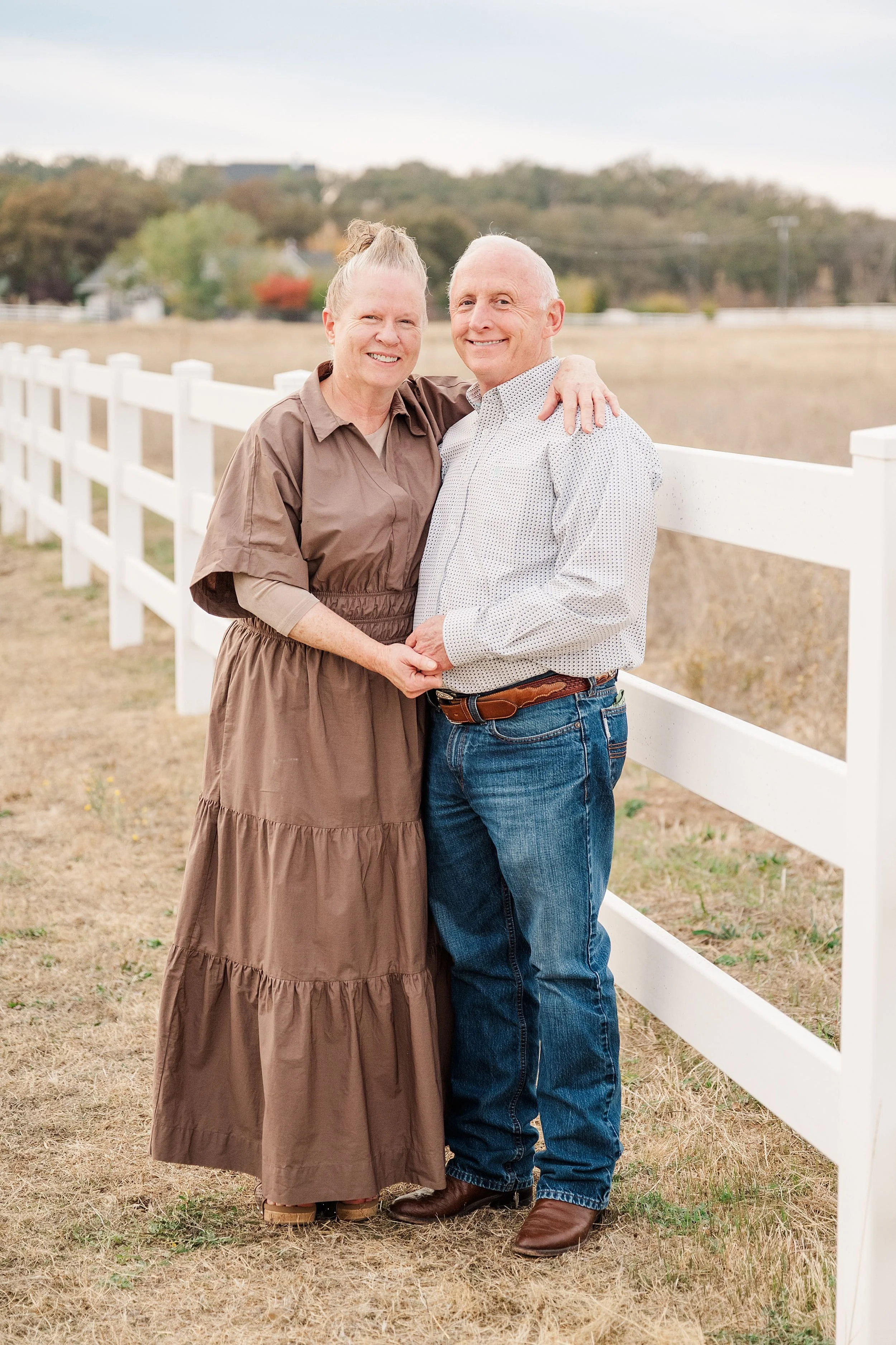 Senior married couple in western attire leaning against white rail fence