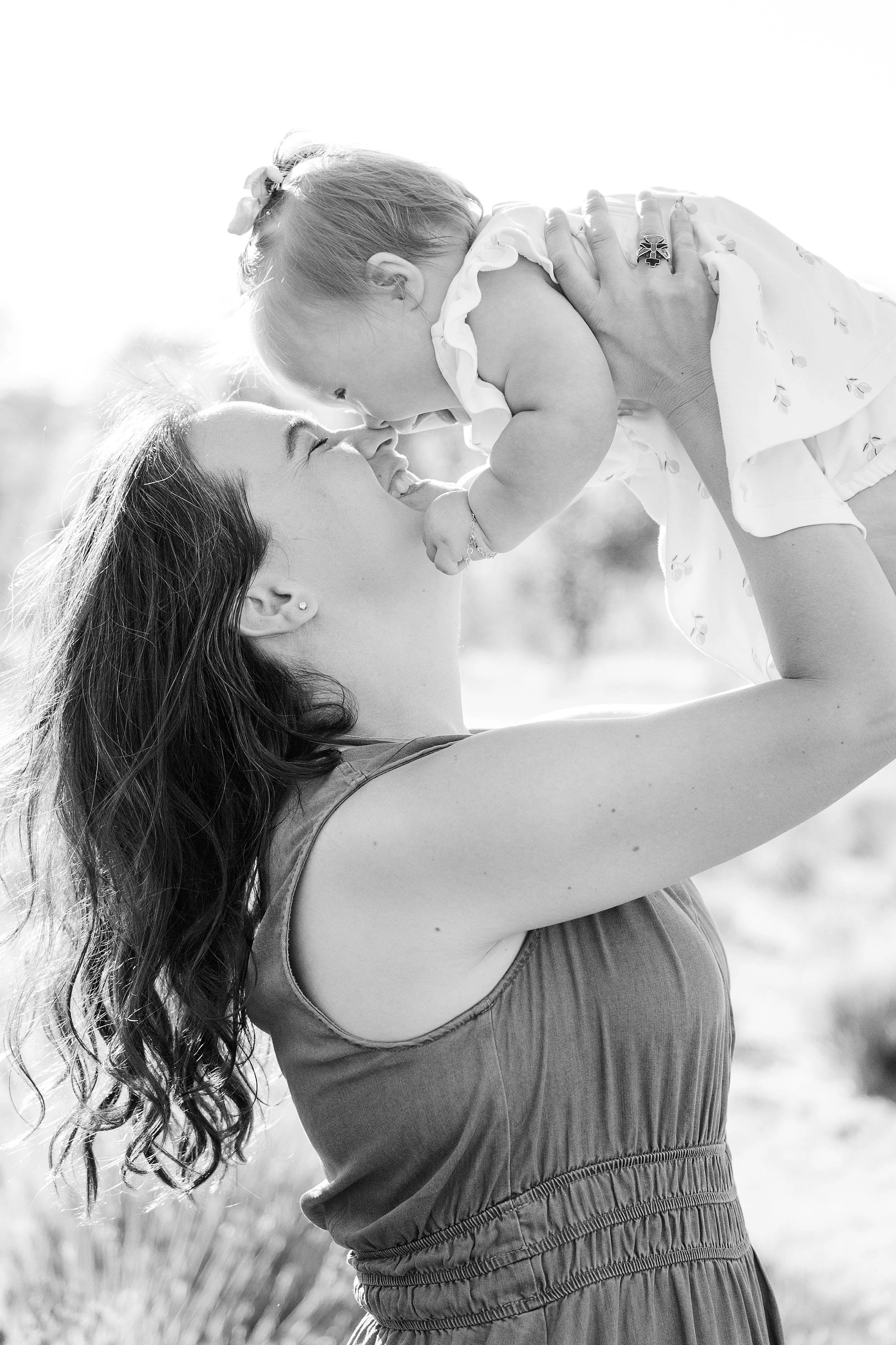 A woman lifts a smiling baby in the air, noses touching, in a sunlit outdoor setting. The scene is joyful and captured in black and white.