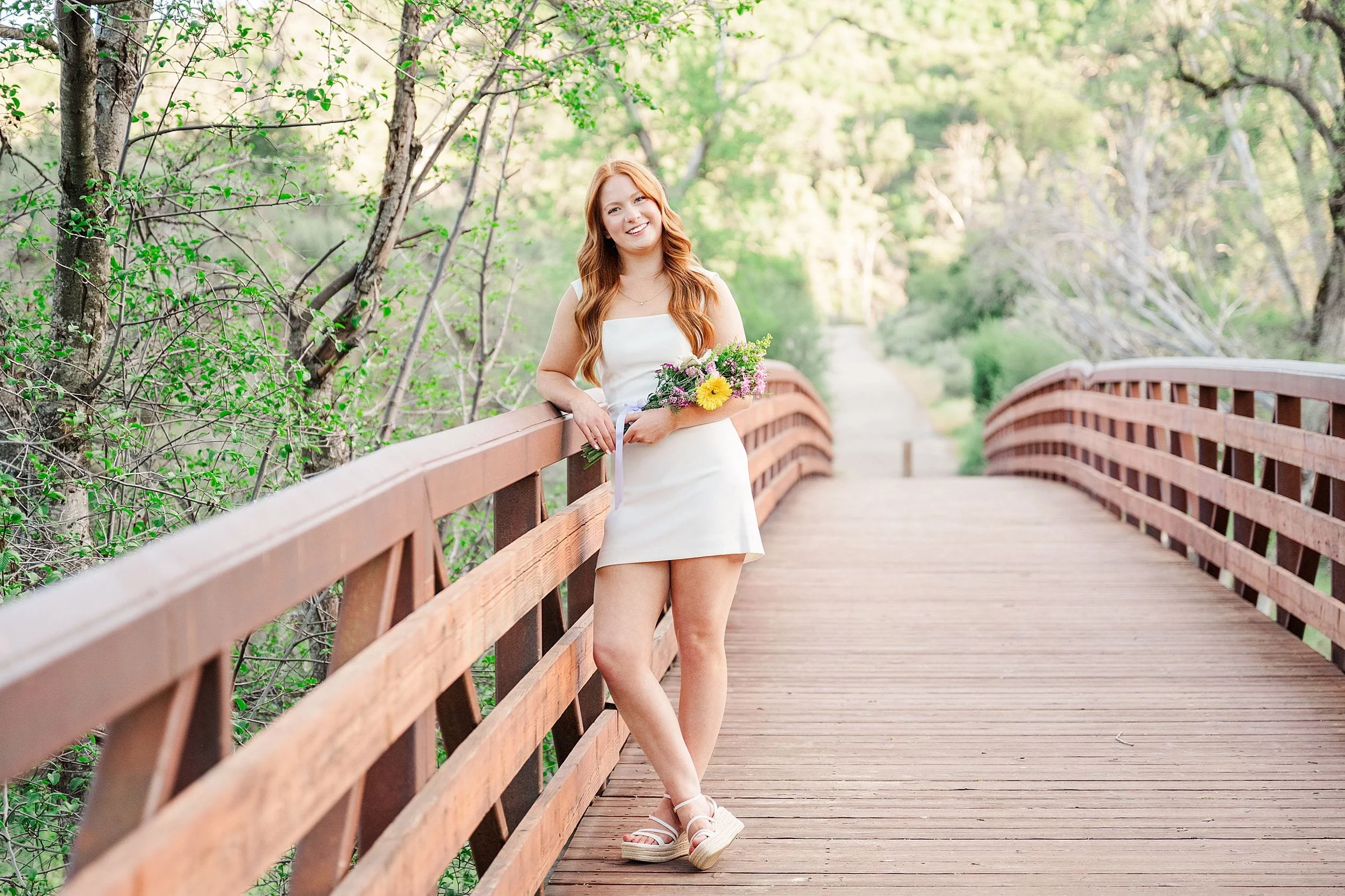 A woman in a white dress stands on a wooden bridge, holding a bouquet of flowers. She smiles gently, surrounded by lush greenery and sunlight.