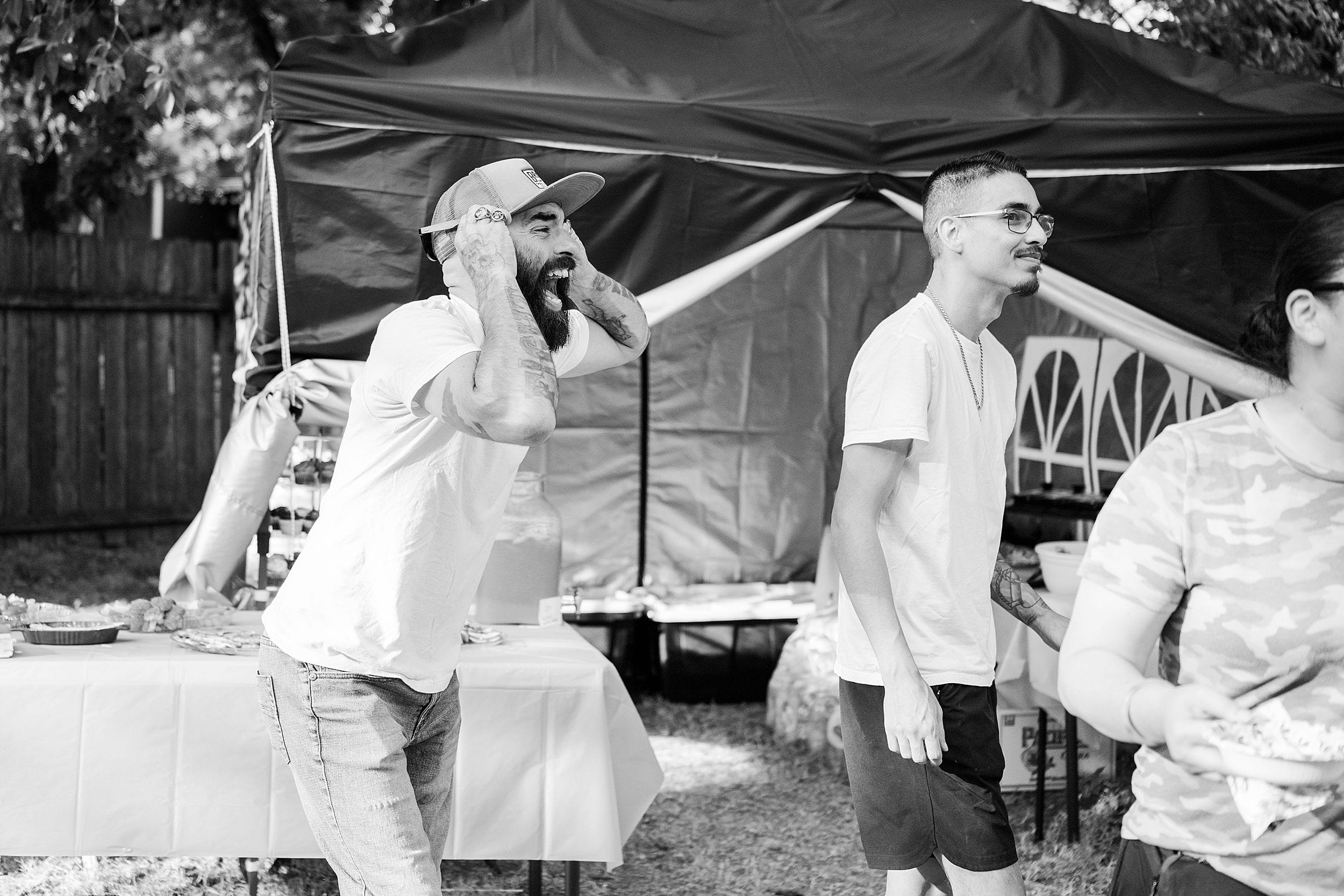 Black-and-white outdoor gathering shows a bearded man in a cap calling out while friends stand beneath a canopy; casual, joyful mood.