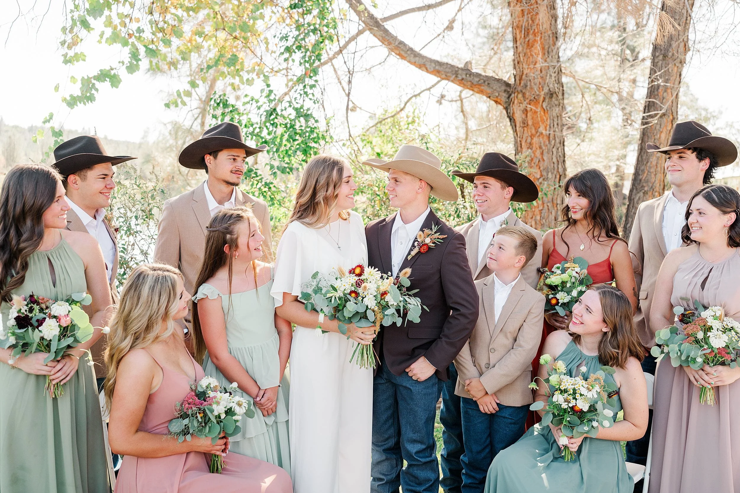 A bride and groom stand smiling beneath a tree, surrounded by bridesmaids and groomsmen in muted pastels, holding bouquets in a warm outdoor wedding portrait.