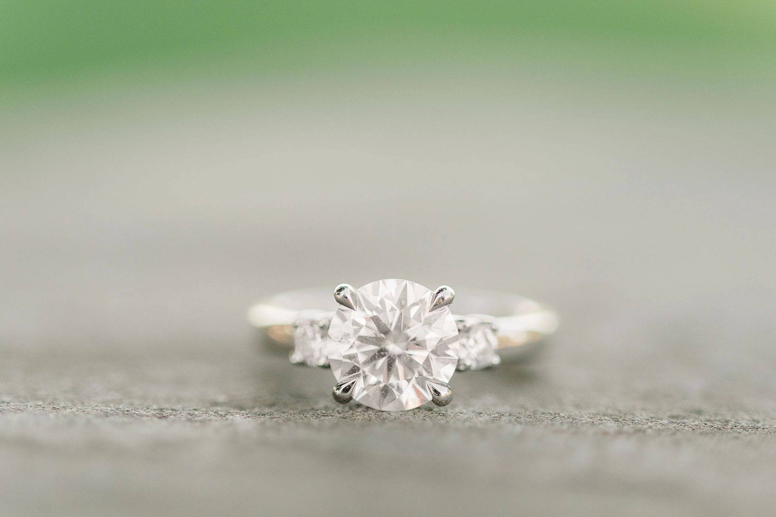A silver engagement ring with a large round diamond centerpiece, flanked by smaller diamonds, sits on a smooth, neutral surface against a blurred green background.