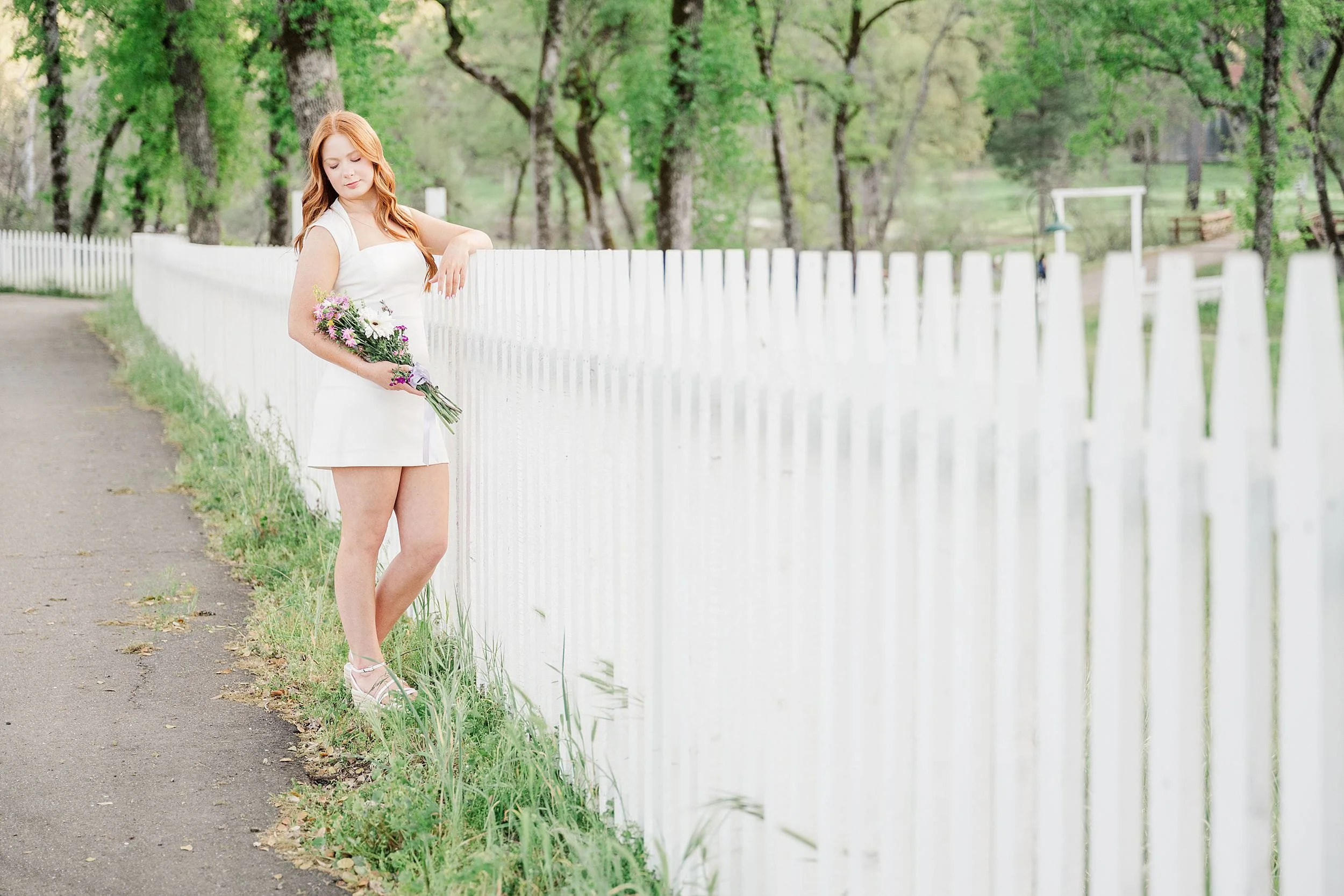 A woman in a white dress stands by a white picket fence, holding a bouquet of flowers. The scene is serene, with lush green trees lining the path.
