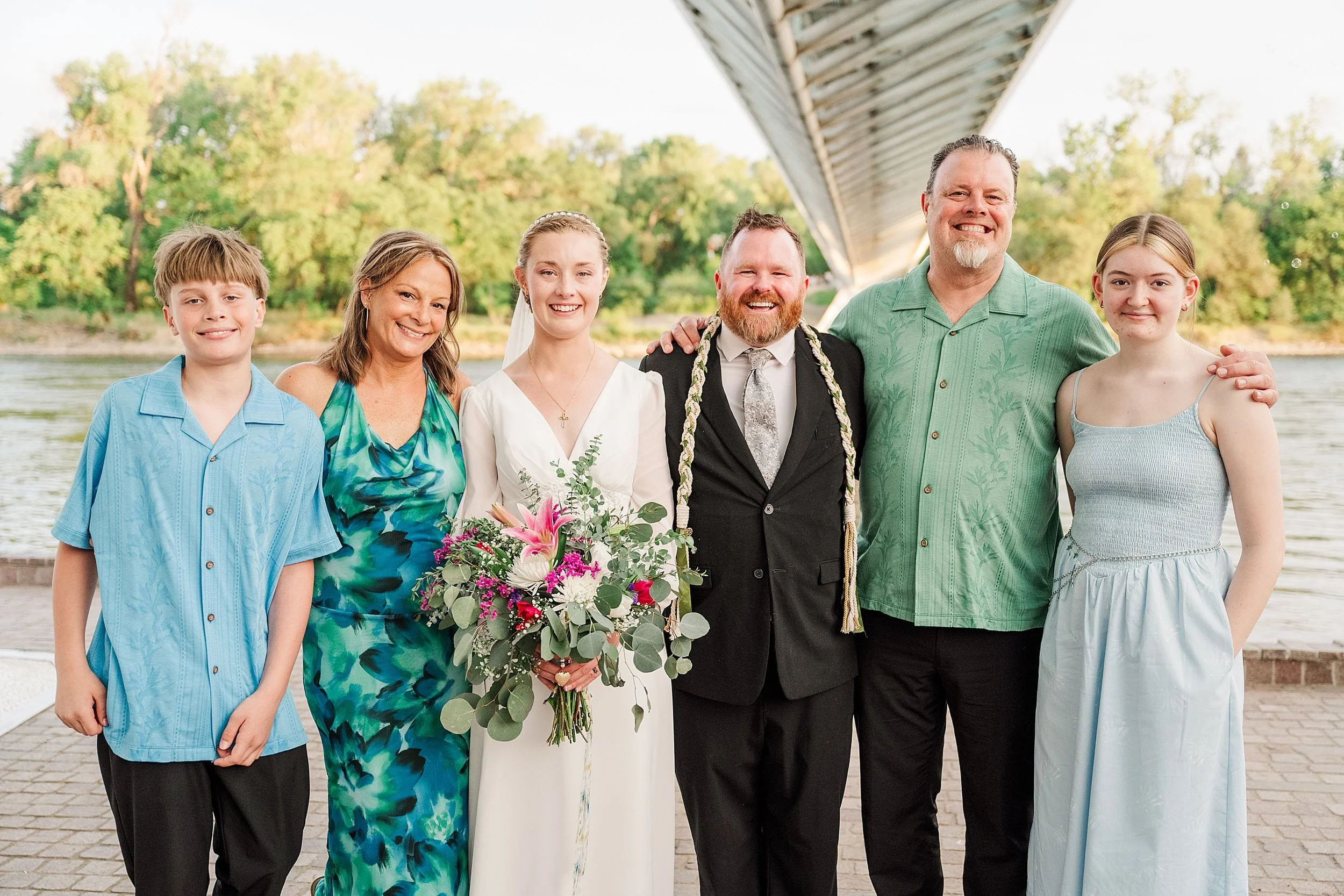 A joyful wedding group photo under a bridge by a river. The bride holds a colorful bouquet, and everyone smiles in summery attire against a lush green backdrop.
