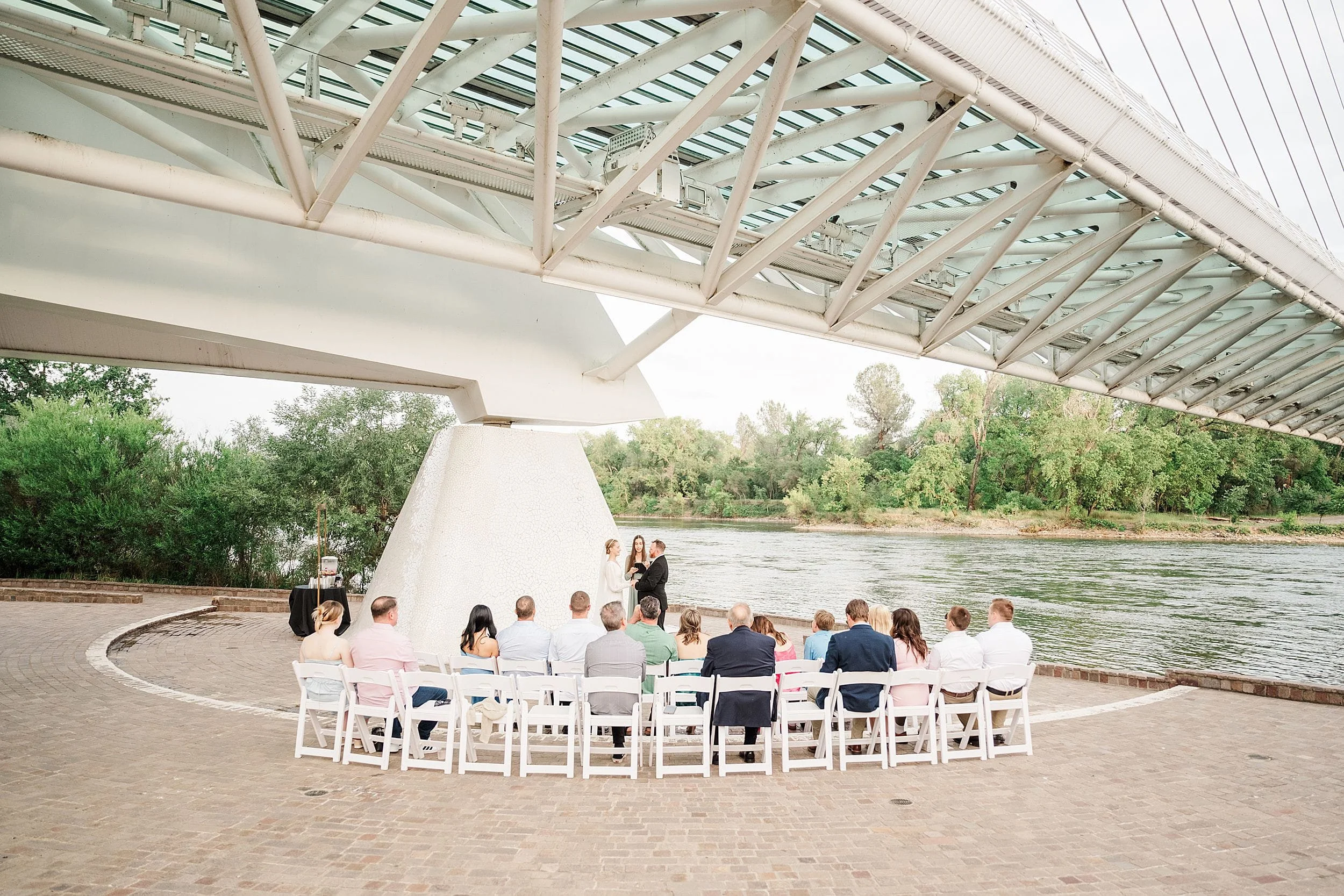 A small group attends an outdoor wedding under a modern white bridge by a river. The atmosphere is serene, with lush greenery in the background.