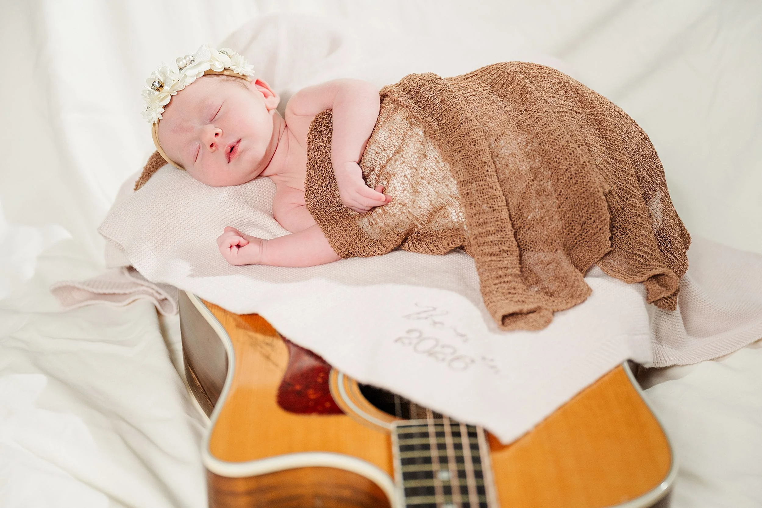 A newborn baby sleeps on a guitar, wrapped in a brown knit blanket, with a floral headband. The scene conveys tranquility and warmth.