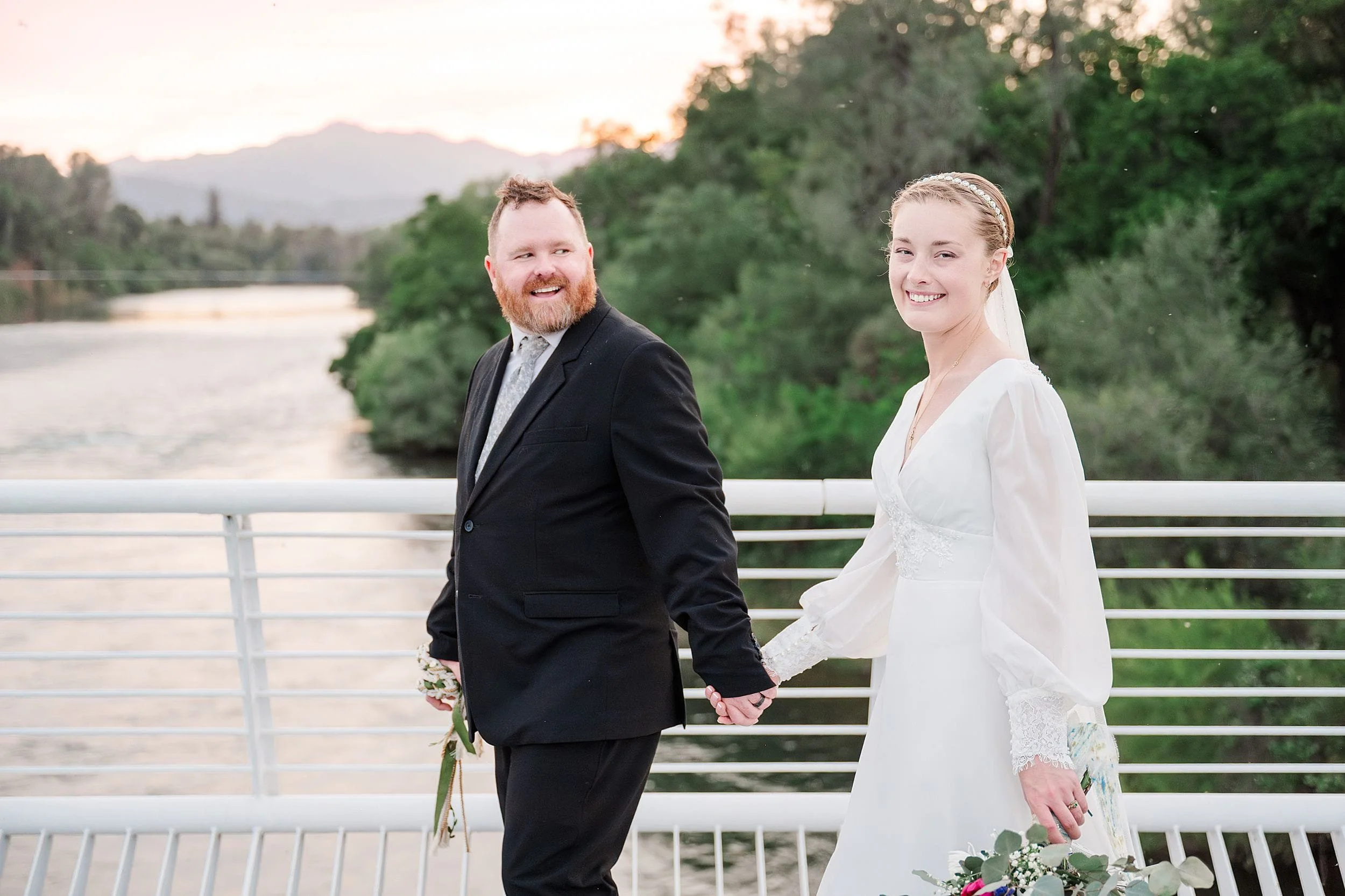 A couple in wedding attire, holding hands and smiling, walk on a bridge near a river with lush greenery. The sky is softly lit by a sunset.