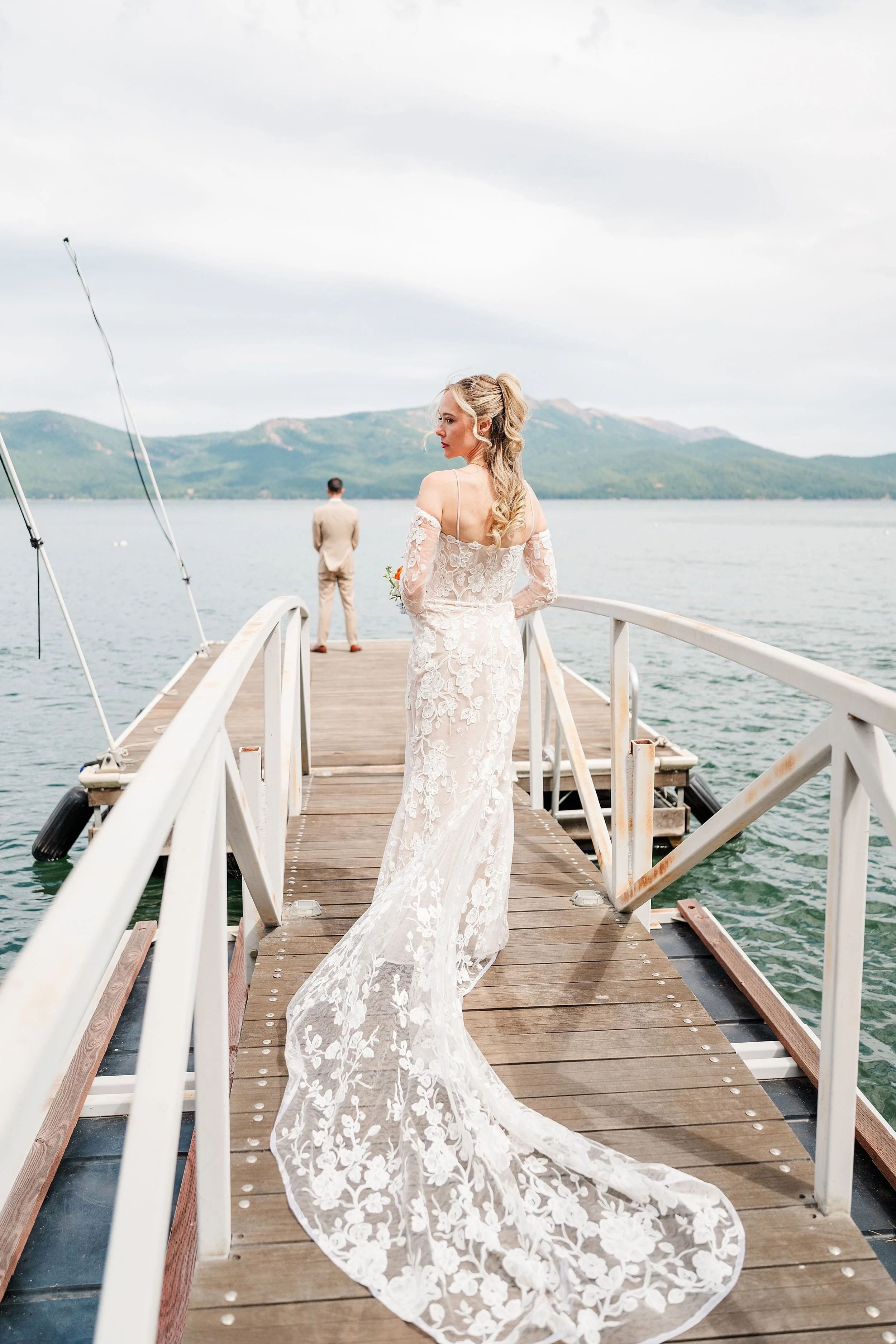 Bride in a lace wedding gown walks along a wooden lakeside pier, her train trailing behind, with mountains, water, and groom ahead.