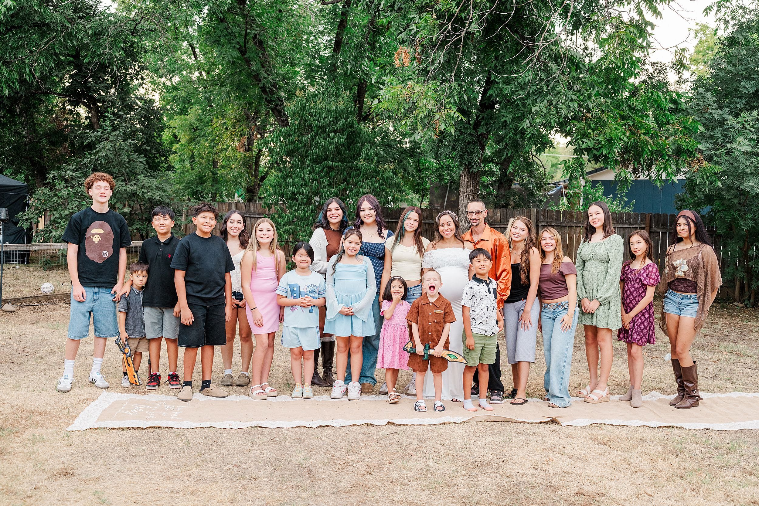 Large multigenerational group poses outdoors beneath leafy trees, smiling in casual summer clothing during a warm, cheerful gathering.