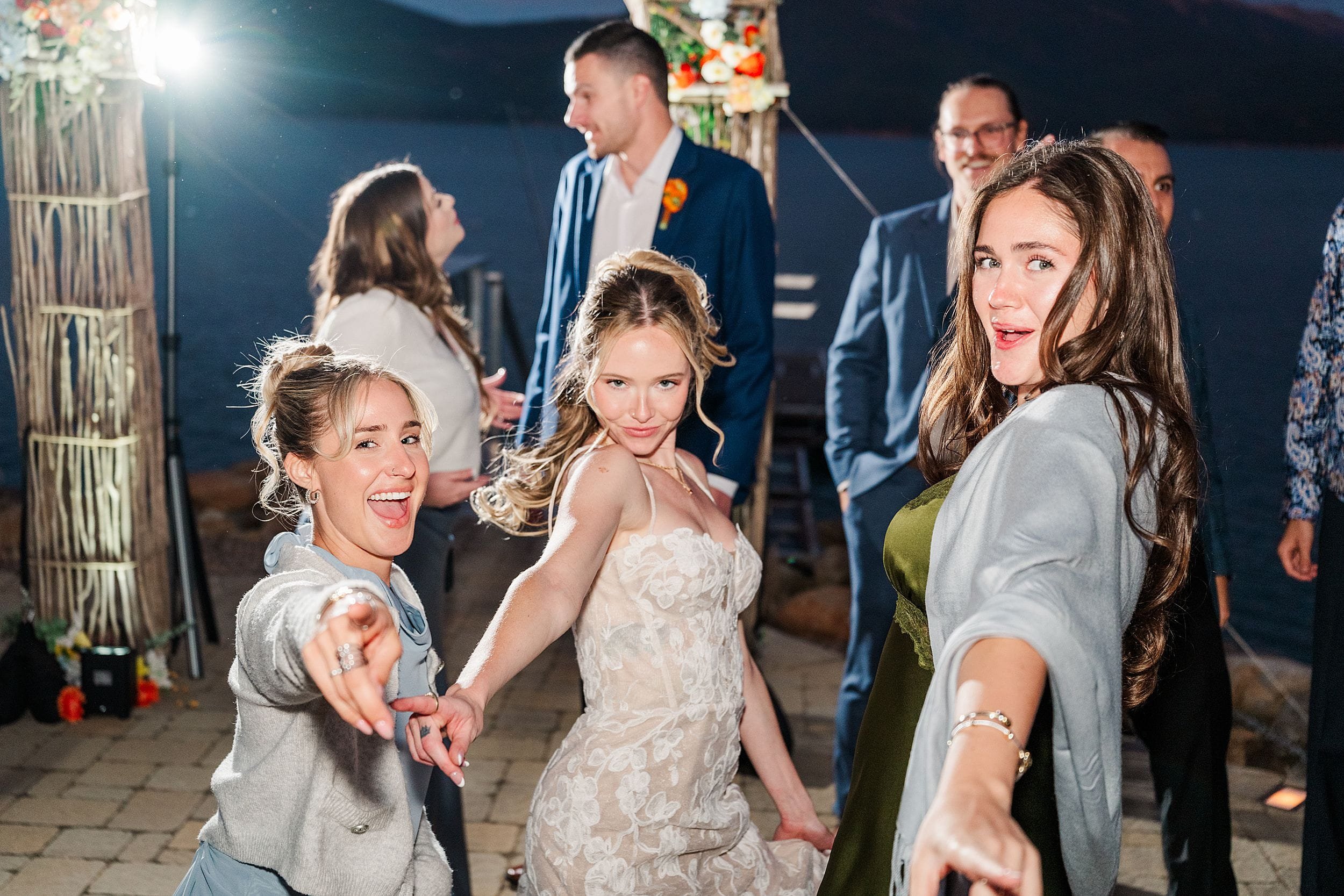 Joyful bride and wedding guests dance outdoors at night, smiling and reaching toward the camera under string lights in a festive setting.