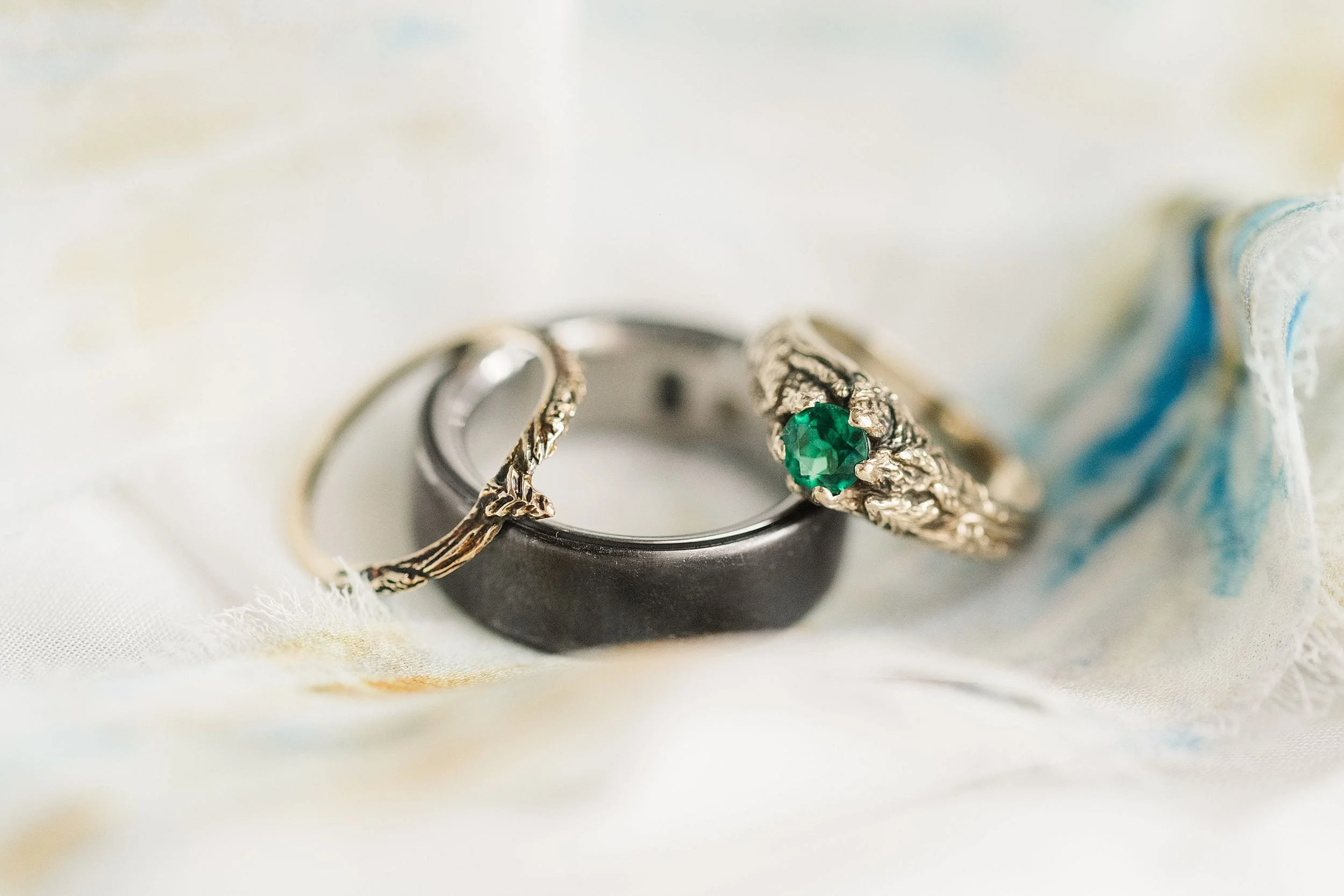 Three rings on a soft, colorful fabric: a simple silver band, a delicate gold ring, and an ornate gold ring with a green gemstone, conveying elegance.