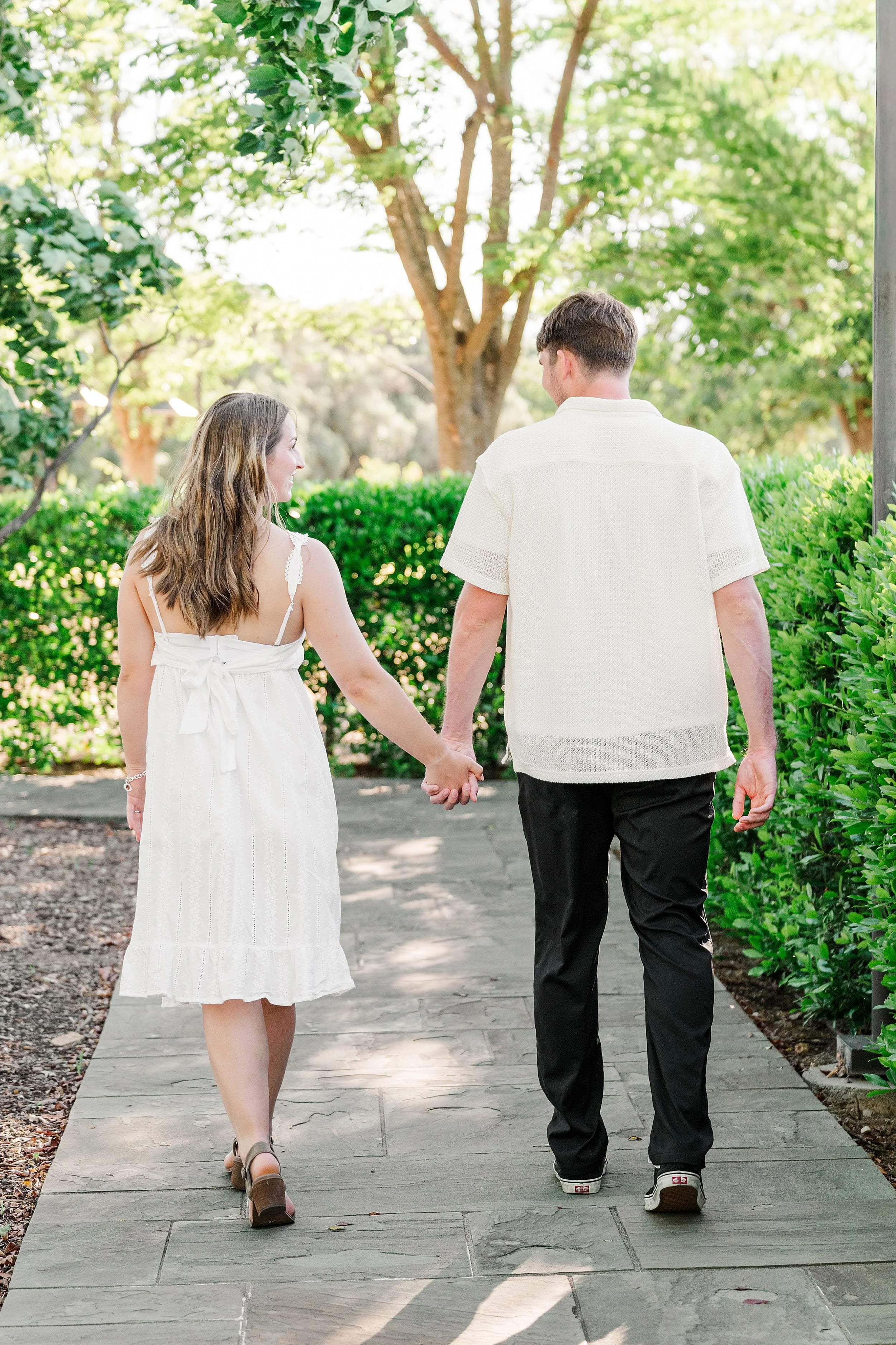 A couple walks hand in hand down a garden path, surrounded by lush greenery on a sunny day. They wear light, summery outfits, creating a serene, romantic vibe.