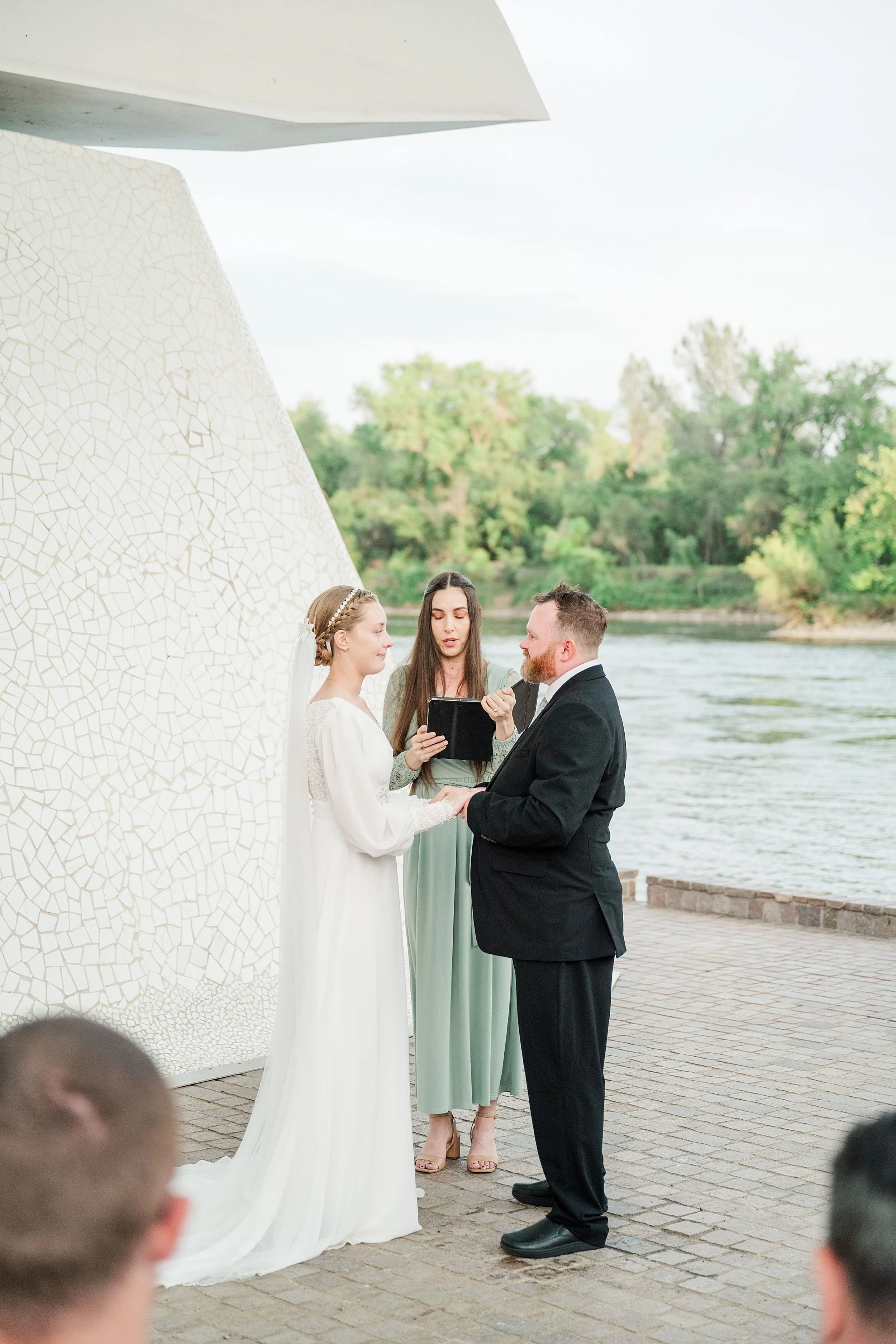 A couple stands facing each other, holding hands, during an outdoor wedding ceremony by a river; the officiant reads vows, creating a serene atmosphere.