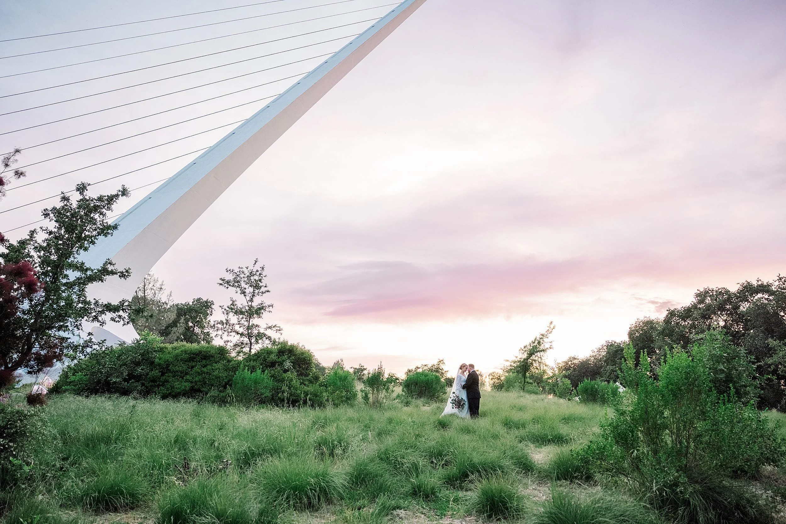 A couple stands on a grassy field at sunset, embracing with a modern cable-stayed bridge to the left. The sky glows pink and purple, conveying tranquility.