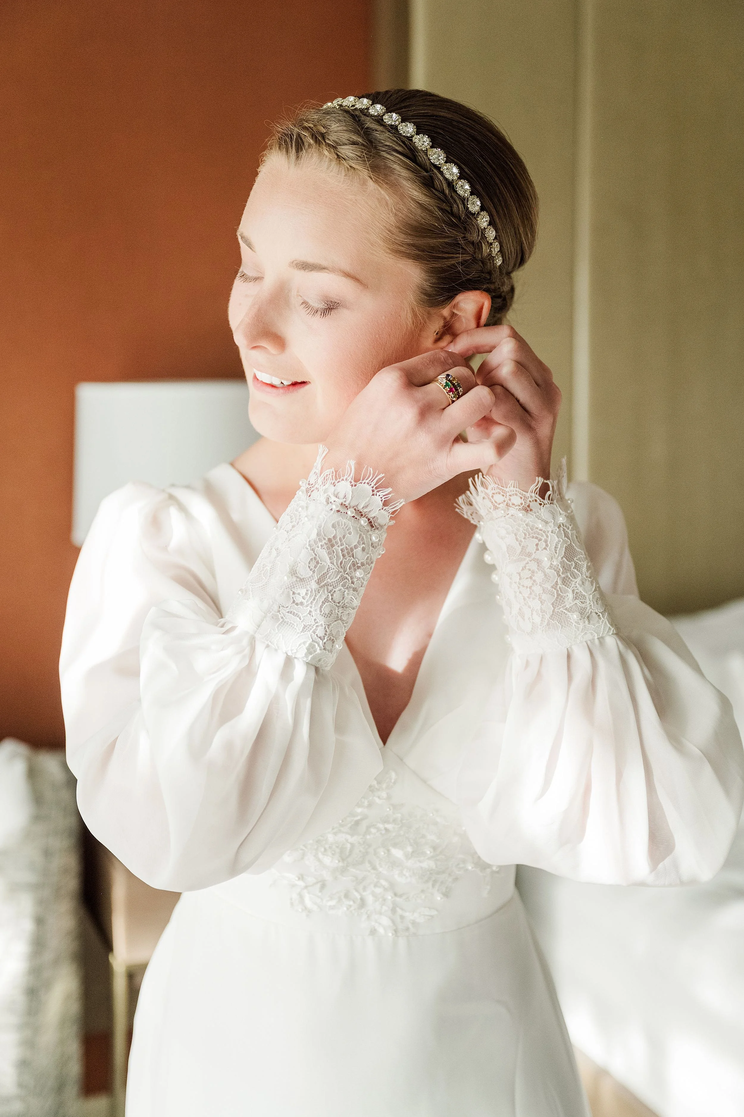 A bride in a white gown adjusts her earring, eyes closed with a serene smile. Her dress features lace details and she wears a bejeweled headband.